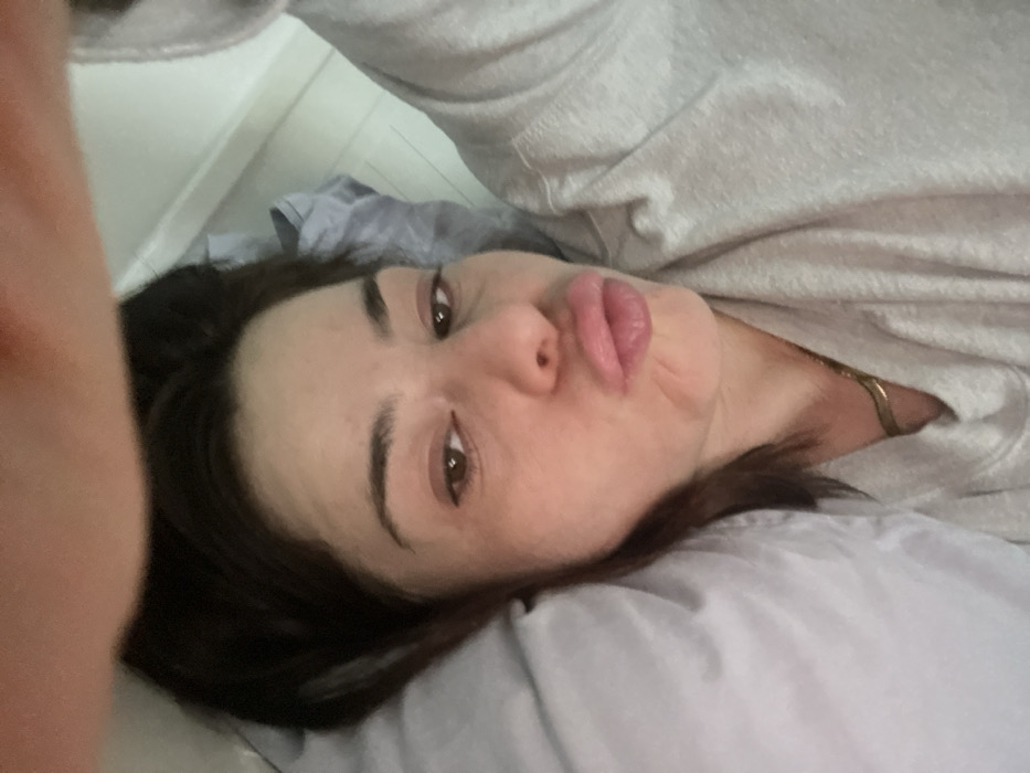A woman lies on a pillow, presumably in bed, wearing a light gray hoodie. She has brown eyes and dark hair. Her lips are pursed in a pout. A gold chain is visible at her neck. The background is a mix of gray and white. The image is taken at an angle, giving a slightly distorted perspective.