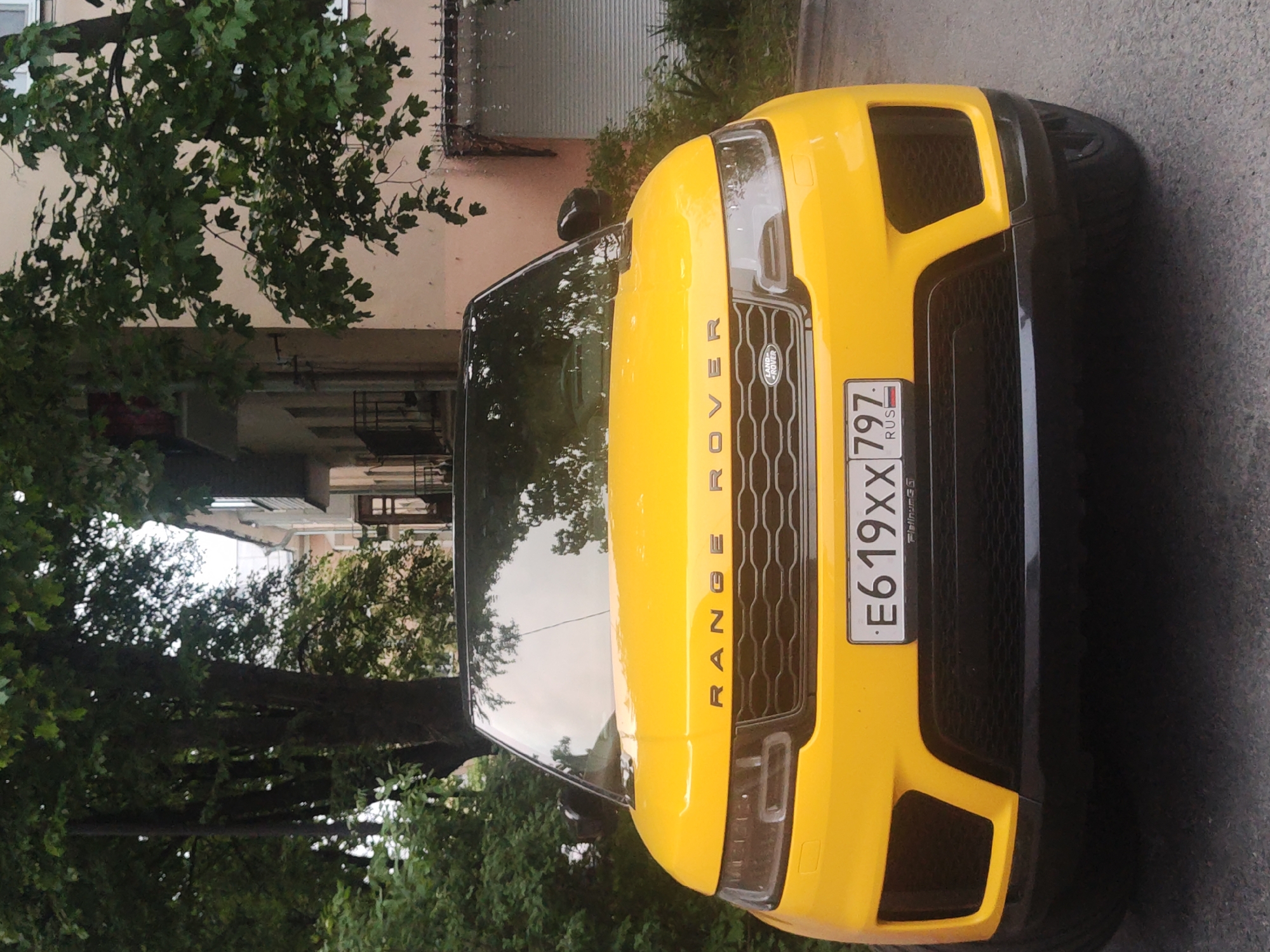 A yellow Range Rover is parked on an asphalt surface in Pushkin, Russia. The car faces forward, revealing its front grille and headlights. The Russian license plate reads "E619XX797". Trees and a multi-story building are visible in the background. The photograph is taken at an eye-level perspective, likely on a street in the city.