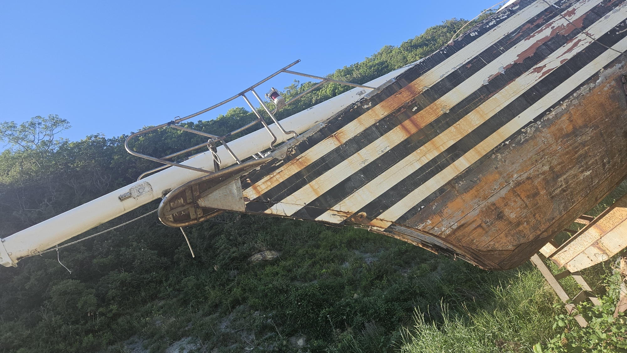 An abandoned, weathered wooden boat with black and white diagonal stripes sits on land against a backdrop of green foliage and a clear blue sky. The boat's hull shows significant signs of age, with peeling paint and rust. A tall, white mast extends upwards from the boat. The scene appears to be located outdoors, possibly in a coastal or rural area, given the vegetation and the presence of a boat. The boat is resting on wooden supports.