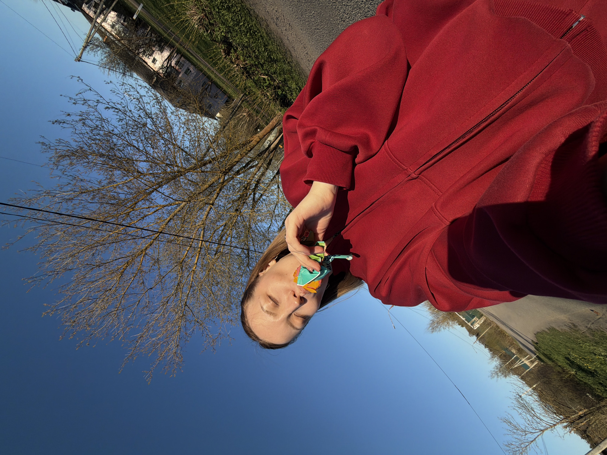 A medium shot depicts a person in a red hooded sweatshirt with their eyes closed, holding a multi-colored, origami-like object near their mouth. The background features a clear blue sky, the bare branches of trees, and a paved road. Distant buildings and greenery line the horizon. Based on the available information, the location is Reshetnikovo, Russia.