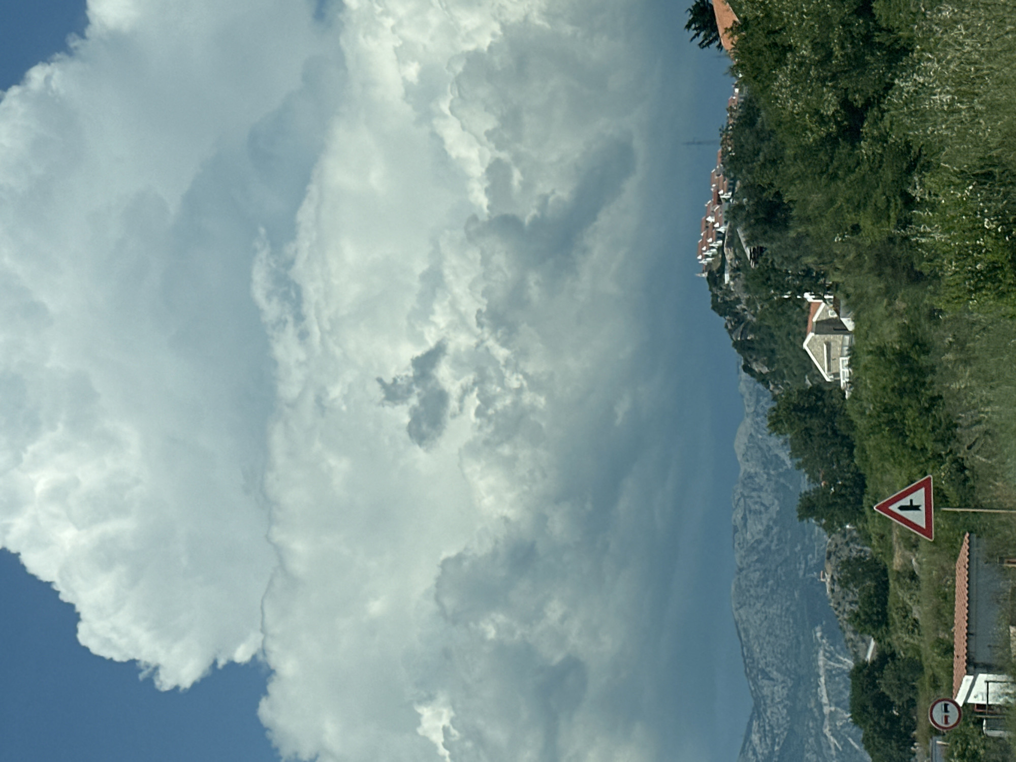 This image captures a scene in Blizikuće, Montenegro, under a dramatic sky filled with large, billowing white and gray clouds. The clouds occupy the upper two-thirds of the frame, with the lower portion featuring a landscape. In the mid-ground, a hillside is covered with lush green trees and vegetation. Several houses with red-tiled roofs are clustered on the crest of this hill, forming a small settlement. To the left of the hill, a building with a red roof is visible. In the foreground, a traffic sign with a red triangle and a black intersection symbol stands on a pole. Another circular traffic sign with a red border and a horizontal bar is partially visible to the far left. The background displays a range of rugged, grey mountains. The overall impression is one of a coastal mountainous region with a developing settlement, under an imposing and overcast sky.