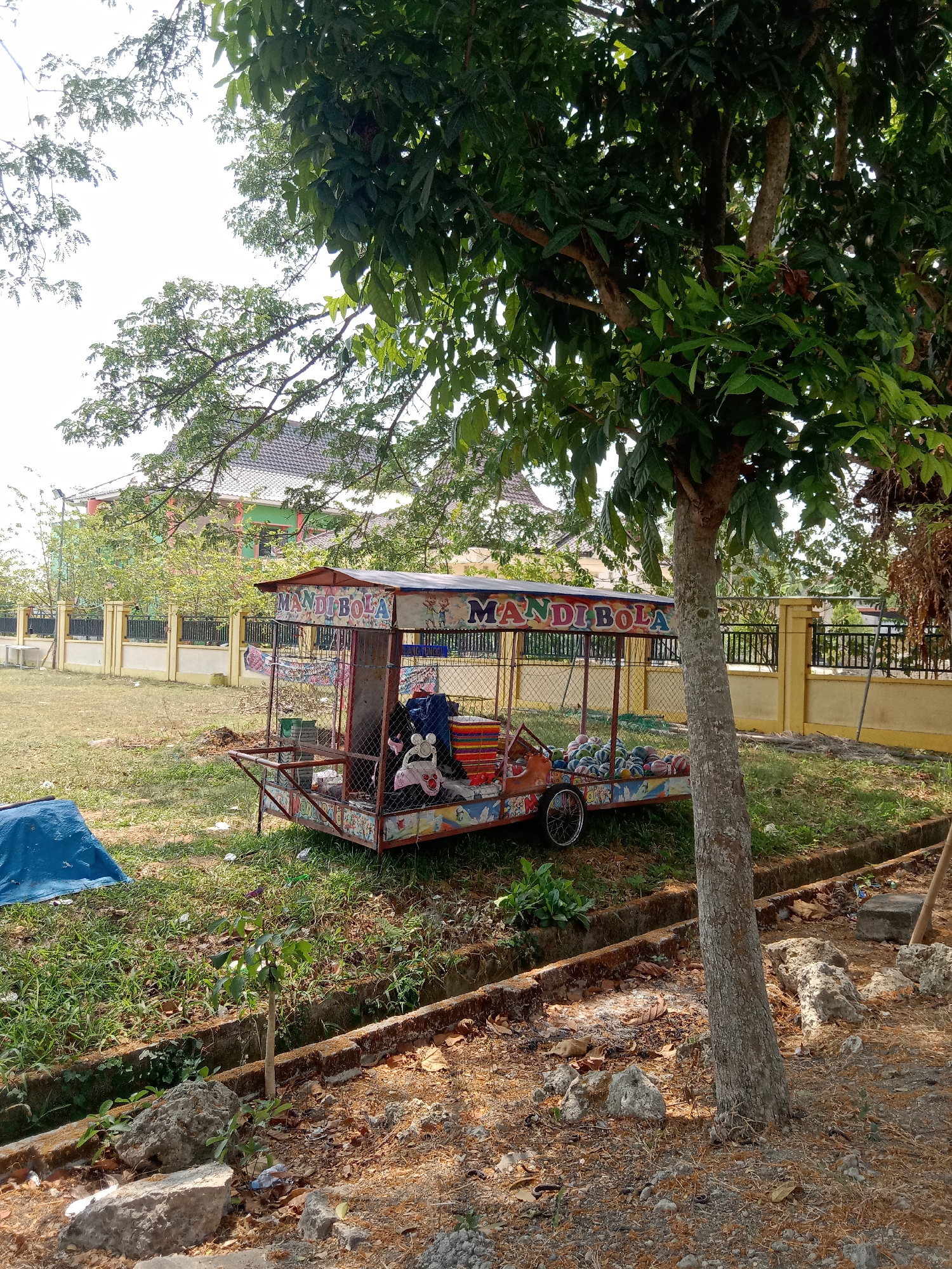 The image depicts a mobile ball pit attraction, labeled "MANDI BOLA," parked on grassy terrain in Kenep, Indonesia. The structure, which appears to be on wheels, is colorful and decorated with cartoon characters. It is filled with numerous balls of various colors. The background shows a residential area with houses and a fence. A large tree with dense foliage is prominently positioned in the foreground, casting shadows on the ground. Other elements visible include a blue tarp, some rocks, and a drainage ditch. The scene suggests an outdoor recreational area, possibly a park or public space, during daytime.