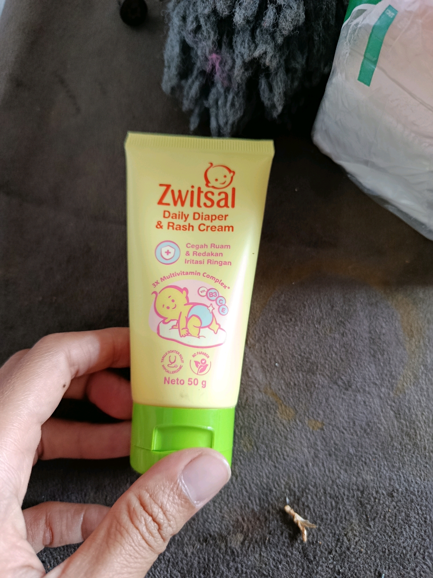 An overhead shot shows a person's hand holding a small tube of "Zwitsal Daily Diaper & Rash Cream" which is yellow with a green cap. The tube is positioned vertically and centrally in the frame. The text on the tube is red and white, and it features a cartoon drawing of a baby. Text on the tube reads: "Zwitsal Daily Diaper & Rash Cream", "Cegah Ruam & Redakan Iritasi Ringan", "3X Multivitamin Complex", "TERUJ DOKTER KULT", "HYPOALLERGENIC", and "Neto 50 g", "NO PARABEN". The tube is held by a person's left hand, with the index finger and thumb gripping it. The background consists of a textured, dark brown surface, possibly a fabric. To the upper right of the tube, a white package with a green label is partially visible. In the upper center of the frame, a dark, fuzzy object with some pink threads is present. To the lower right of the tube, a small, light brown insect-like object is visible on the brown surface. The scene is indoors, with no indication of it being in the city of Karang Rejo, Indonesia.