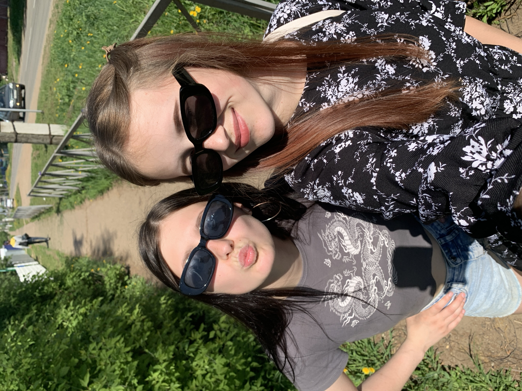 Two young women are posing for a picture outdoors in Фоки, Russia. The woman on the right has long, light brown hair and is wearing a black and white floral print dress and sunglasses. The woman on the left has long, dark hair and is wearing a gray t-shirt with a dragon design, denim shorts, and sunglasses. They are standing next to a metal railing and a sidewalk, with green foliage and yellow flowers visible in the background. A road and some buildings can be seen further in the distance.