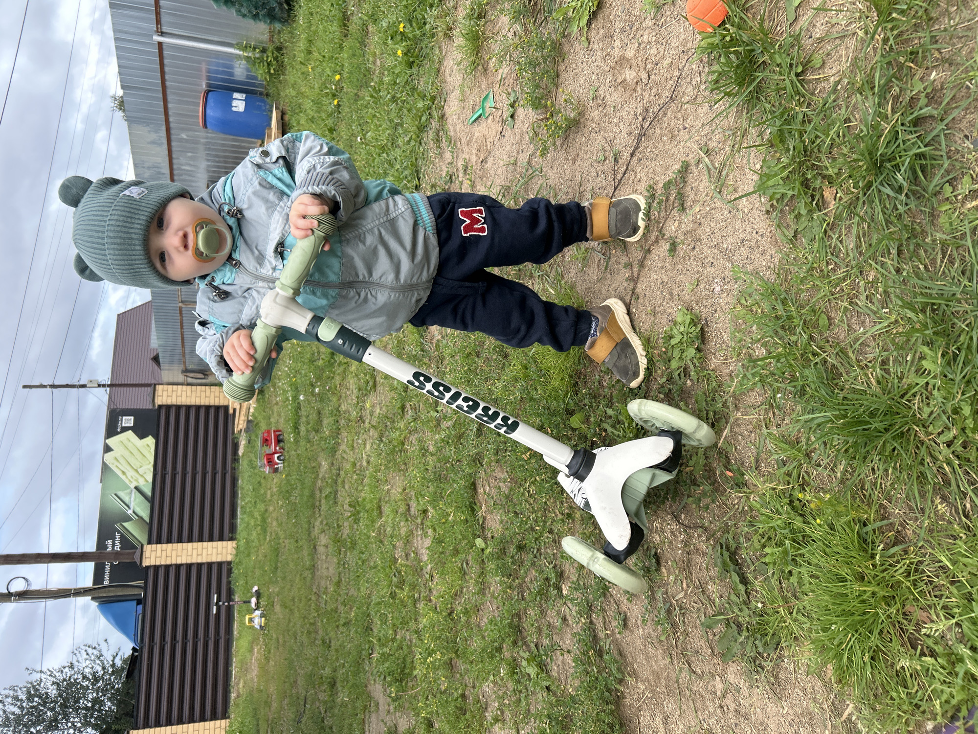 A young child wearing a green beanie with ear details, a grey and teal jacket, and dark sweatpants with a red "M" patch stands on a dirt and grass patch outdoors, holding the handlebars of a three-wheeled scooter. The child has a pacifier in their mouth. The background features a corrugated metal fence, a tall utility pole, a blue barrel on a wooden pallet, and a building with a brown roof. In the distance, a fence with a large advertisement is visible. Scattered toys, including a red toy fire truck and a yellow and white toy scooter, are on the grass in the background. The weather appears overcast. The image is likely taken in a residential area of Chaikovsky, Russia, given the presence of typical Russian fencing and advertising.