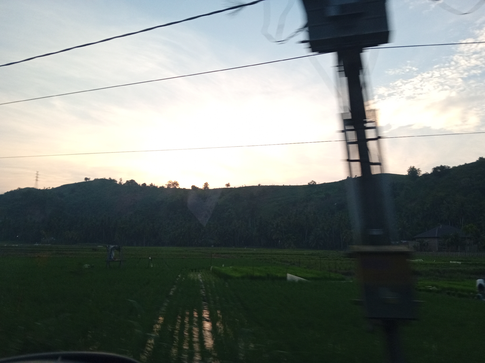 The image captures a rural Indonesian landscape at dusk, likely in Uleejalan, Indonesia. The foreground is dominated by a flooded rice paddy, reflecting the soft light of the setting sun and the sky. The water is disturbed, suggesting movement, possibly from wind or being traversed. Beyond the paddy, a dense, dark green hillside rises, covered in trees, and culminating in a ridge against the horizon. The sky above is a gradient of soft blues and pale oranges, with wispy clouds scattered across it. In the mid-ground, a rudimentary wooden structure, possibly a scarecrow or a small shelter, stands in the paddy. Further in the distance, a cluster of buildings, including a house with a dark roof, is nestled amongst the trees on the hillside. A tall, dark, blurry structure, possibly a utility pole or part of a vehicle, obstructs the right side of the frame, and thin, dark lines, likely power lines, stretch across the upper portion of the image. The overall impression is one of natural beauty and agricultural life in the twilight.