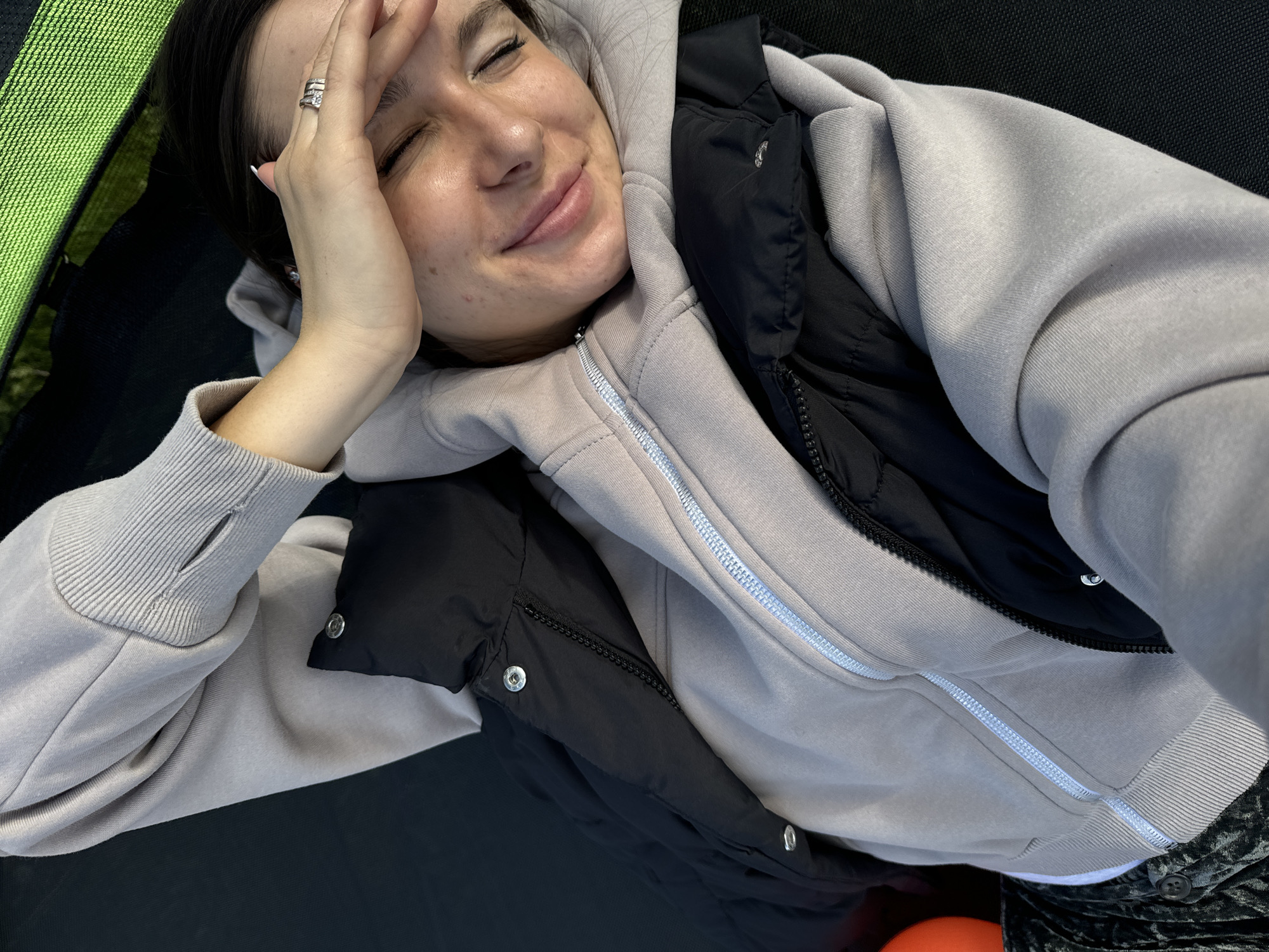 A woman lies on a black trampoline in an outdoor setting, possibly in Vysokinichi, Russia. She is wearing a light gray hooded sweatshirt over a black padded vest. The woman's eyes are closed, and she is smiling with her right hand on her forehead, which has a silver ring on one of the fingers. Green trampoline springs are visible in the upper left.