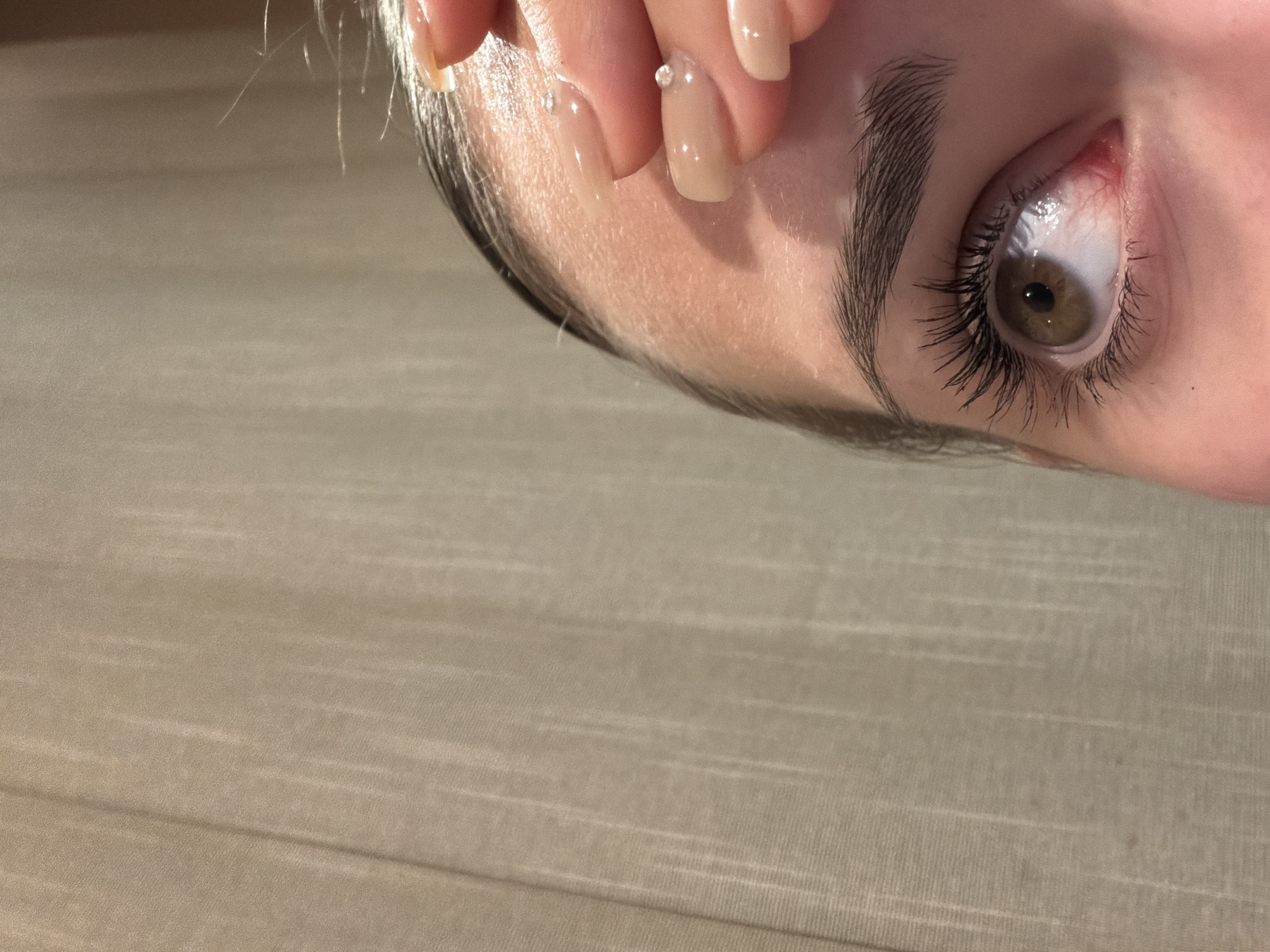 The image depicts a close-up of a woman's face. Her right eye is prominent, with long eyelashes and a brown iris. Above the eye is a well-defined eyebrow. Her forehead is visible, with a few strands of hair along the hairline. Her fingers are partially covering her forehead, and her nails are manicured with a light nude color and small decorative stones. The background appears to be a neutral-colored, textured surface. The location is not discernible from the image alone and cannot be identified as Buhovo, Bulgaria.