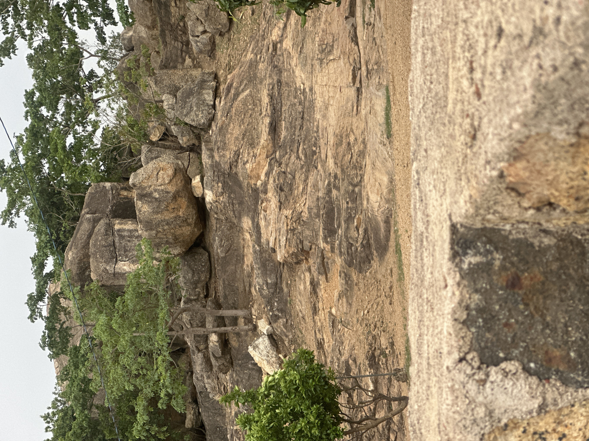 The image depicts a rocky landscape, possibly a cliff or hillside, with trees and vegetation. The terrain is predominantly brown and grey, suggesting rocky or arid conditions. A concrete or stone structure is visible in the foreground. This may be a part of Riyom Rock or Shere Hills, areas surrounding Jos, Nigeria, known for their rock formations. There is no visible presence of people, animals, or any specific activities happening. The scene consists of natural geological features and some elements of the built environment.