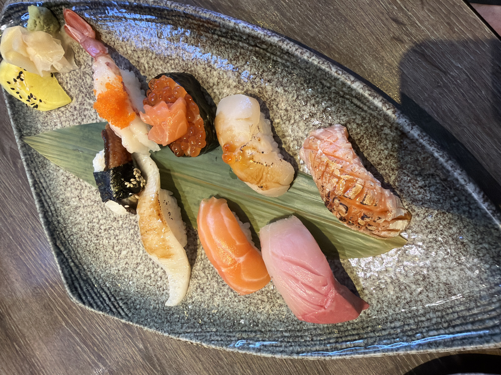 A top-down, close-up shot shows a sushi platter presented on a gray, irregularly shaped plate with a textured surface. The plate rests on a wooden table. A variety of sushi pieces are arranged on the plate, including shrimp sushi topped with orange roe, sushi with salmon roe, seared scallop sushi, grilled salmon sushi, salmon sushi, and tuna sushi. Pickled ginger, wasabi, and a yellow condiment decorated with black sesame seeds are also arranged on the plate. The sushi pieces are placed on a green leaf. The location is บ้านบางรักษ์, Thailand.