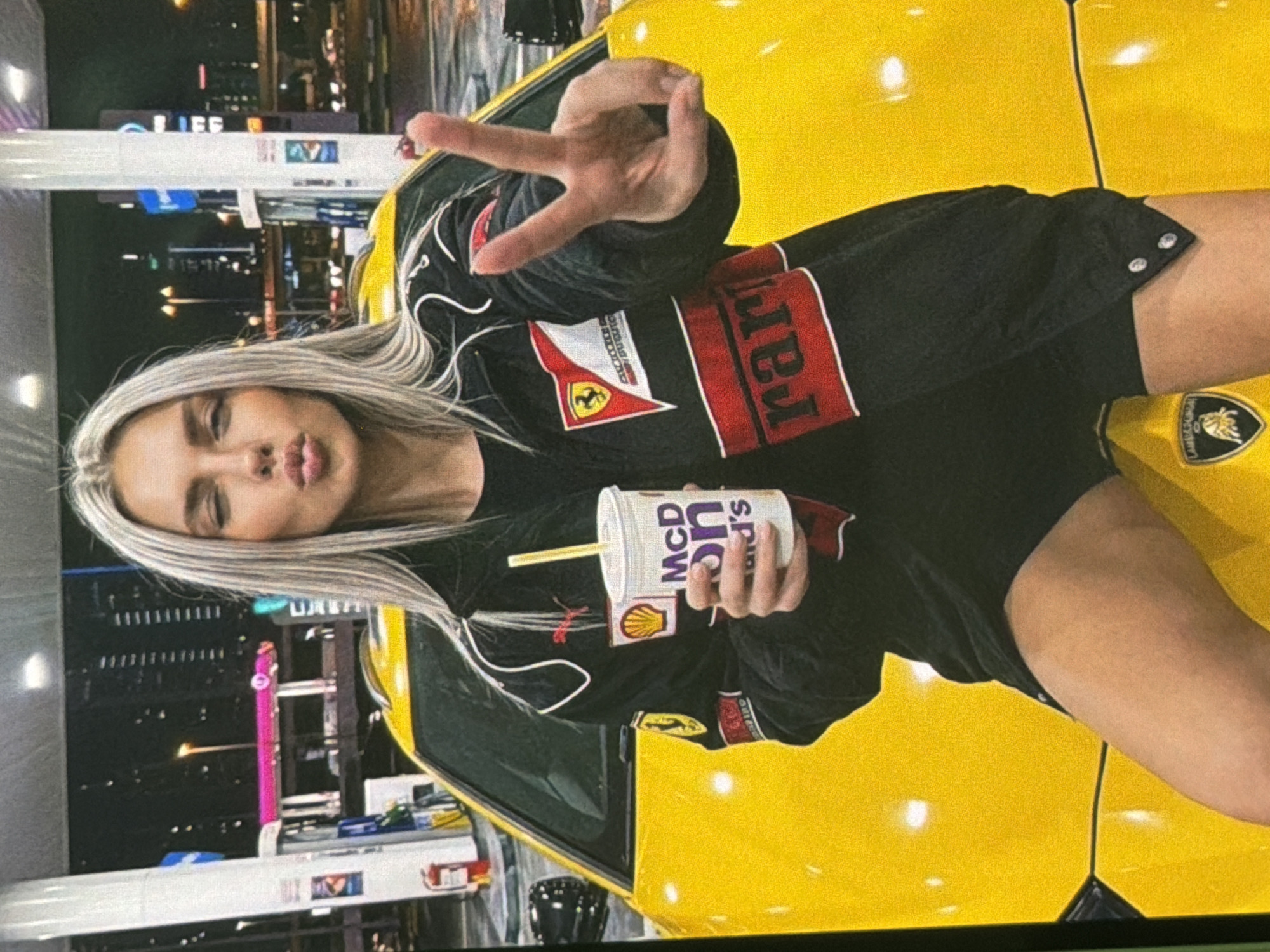 In Bobrovo, Russia, a blonde woman with her lips pursed sits on the hood of a bright yellow Lamborghini at a gas station at night. She is wearing a black racing jacket with red and white accents, featuring logos such as Ferrari and Shell. In her right hand, she holds a McDonald's cup with a straw. Her left hand is raised, forming a peace sign. The background shows gas pumps, city lights, and buildings, indicating an urban environment.