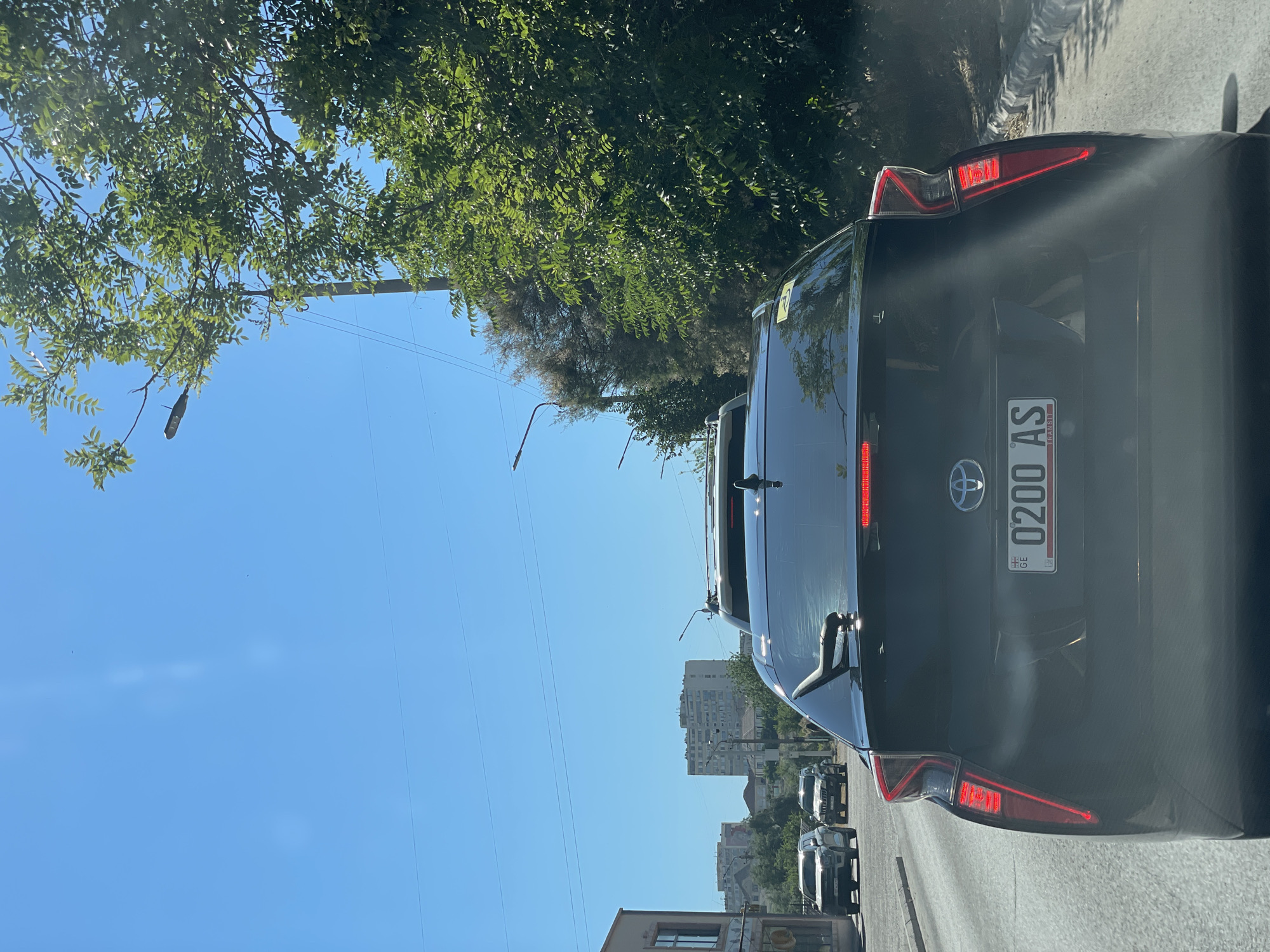A black car is in the foreground, its rear end facing the viewer. The license plate is visible and reads "0200 AS GE". The car is in traffic, with other vehicles ahead and behind it. Tall green trees with lush foliage are on the left side of the image, casting shadows on the road. The sky is a clear, bright blue, suggesting a sunny day. In the distance, several multi-story buildings are visible, hinting at an urban or suburban setting. The perspective of the photo, taken from inside another vehicle, implies the viewer is also in traffic. The scene appears to be set in Aktau, Kazakhstan, during daylight hours with pleasant weather. There are no people clearly visible. The overall mood is neutral, characteristic of everyday traffic.