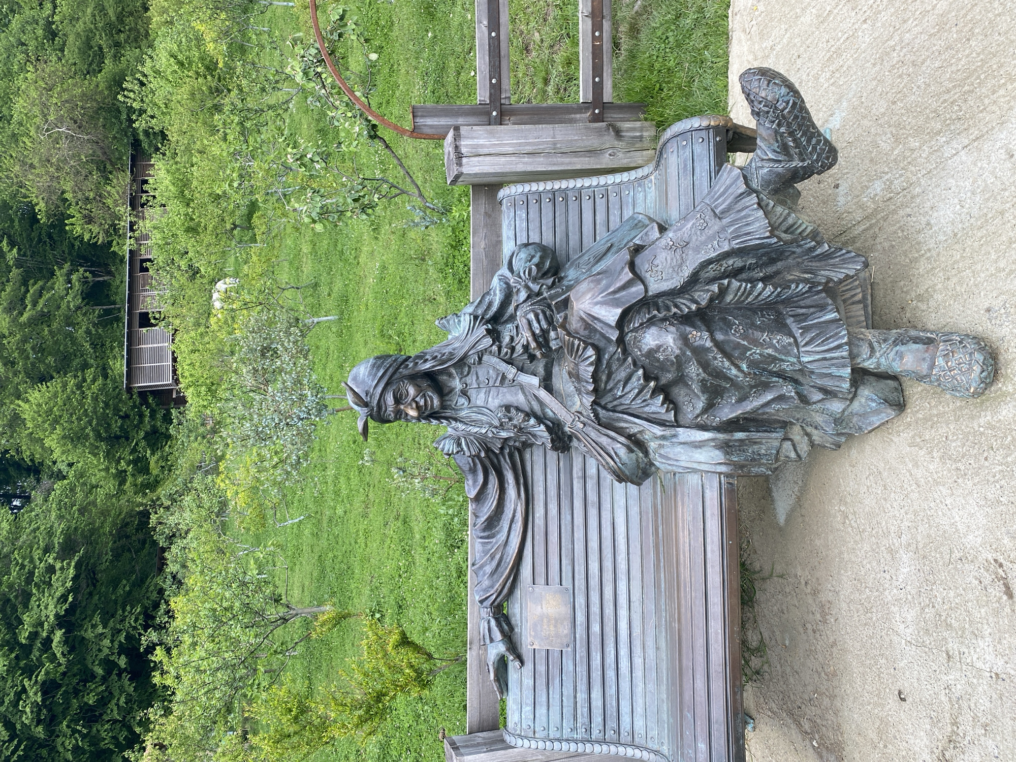 The image depicts a bronze statue of a man sitting on a bench in an outdoor setting. The statue appears to be historical in nature, as the man is wearing period clothing and footwear. He has a hat on and appears to be holding a small child or doll. The bench is also made of bronze and has a plaque attached to it. The background shows a green lawn, trees, and a building with slatted walls. Based on the prompt, this statue may be located in the city of Прогресс, Russia. The statue is sitting on a concrete slab next to green grass and shrubbery. A wooden structure appears in the background.