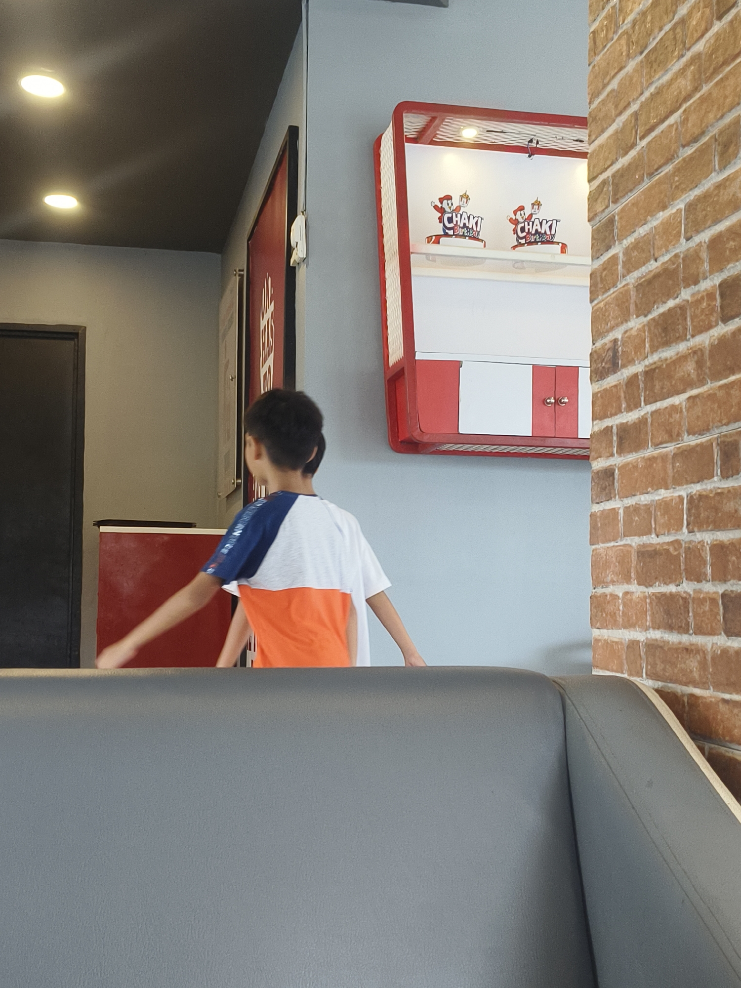 The image shows the interior of what appears to be a fast-food restaurant. A young boy, viewed from behind, is wearing a t-shirt with white, blue, and orange panels. He is positioned in the foreground, with his right arm extended forward. Behind him, a red and white display case is mounted on a light blue wall. This display case contains two identical logos that read "CHAKI Birthday" with cartoon characters. To the right of the display case, a brick wall is visible. In the background, a dark doorway and a red counter are also present. The ceiling is dark gray with two circular light fixtures. The image is a still from a video, suggesting an indoor scene, possibly a public space like a restaurant.