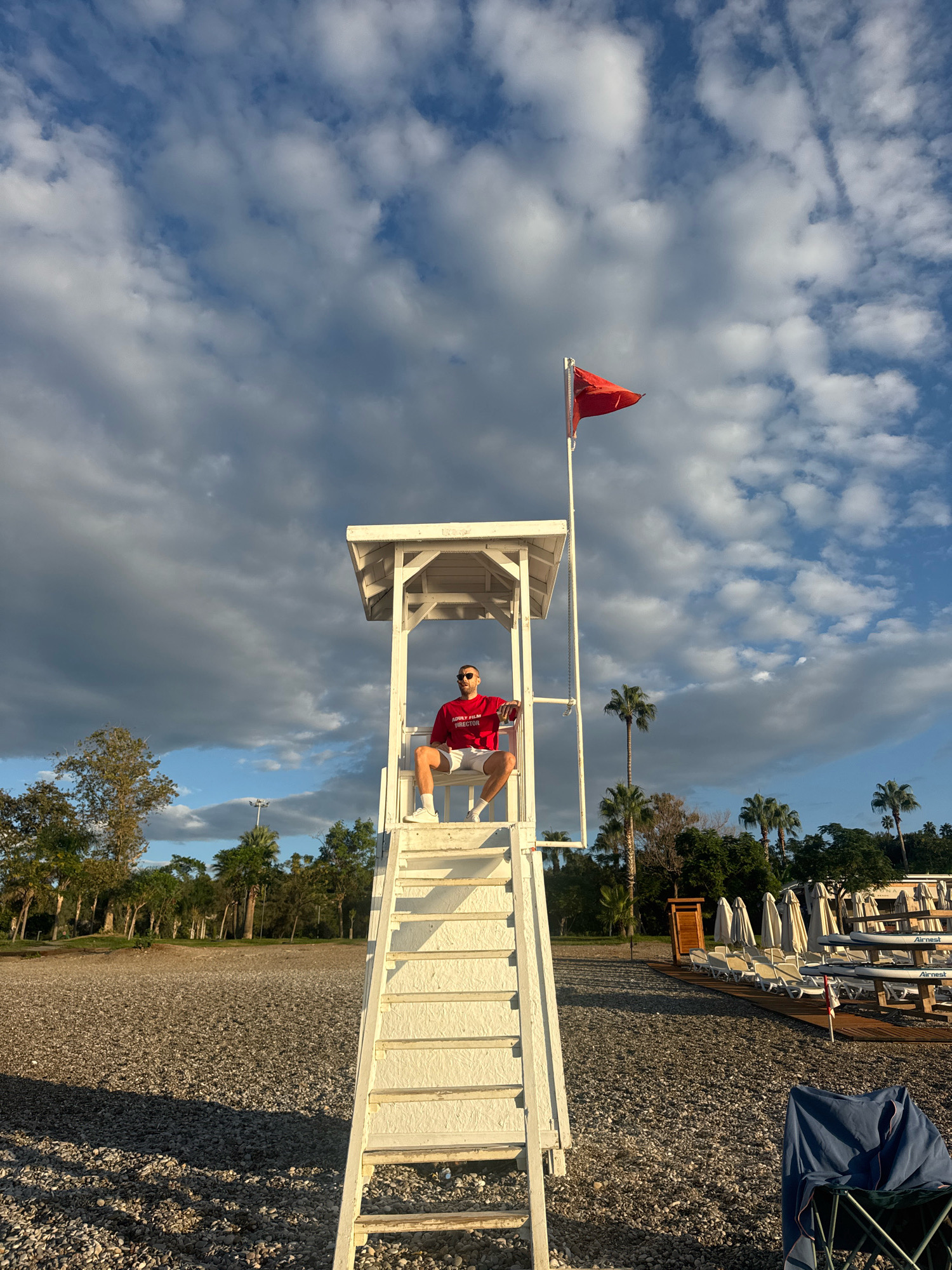 A lone man, wearing a red t-shirt and white shorts, sits on a white lifeguard chair with a red flag flying from the top. He is positioned in the middle of a gravel beach, which stretches out towards a line of trees and palm trees in the background. The sky is partly cloudy with dramatic, dark clouds on the left and brighter, white clouds on the right. The sun is low in the sky, casting long shadows. Beach chairs and umbrellas are visible further down the beach. The scene appears to be on the coast of Antalya, Turkey, likely at a resort or public beach.