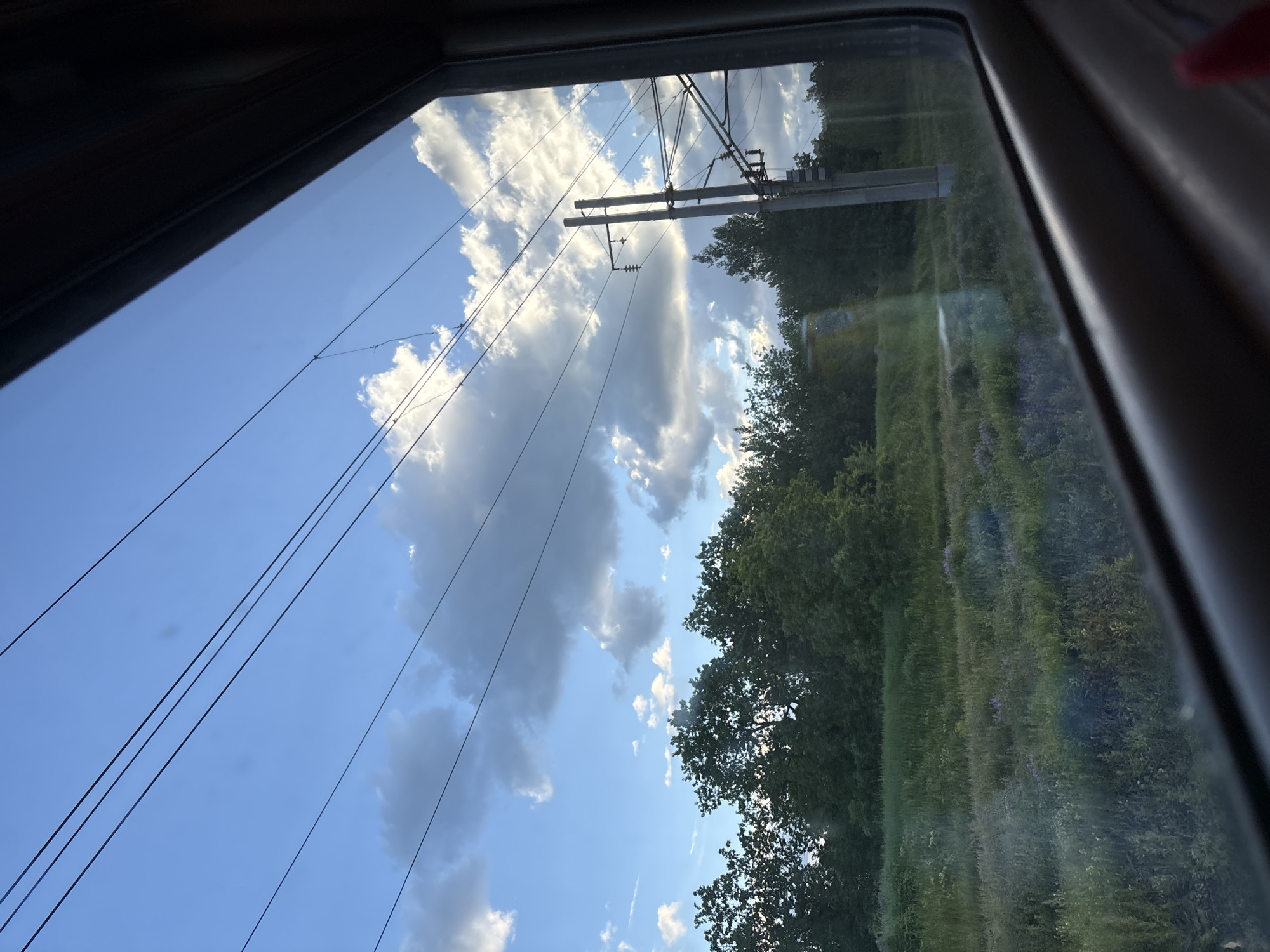 A view from a train window shows a bright blue sky partially obscured by large, white cumulus clouds. Power lines stretch diagonally across the sky, and a utility pole is visible near the center of the frame. Lush green vegetation, including trees and grassy fields, occupies the lower right portion of the view. The image is taken through the train window, which is slightly reflective and shows a portion of the train's interior. The location is speculated to be Karamysh, Russia.