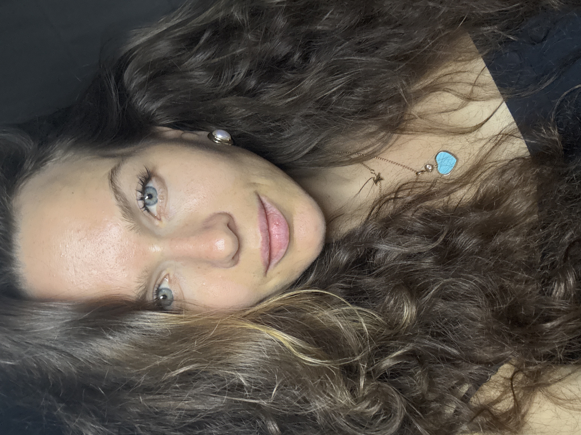 A close-up portrait features a young woman lying down, with her head positioned on the left side of the frame and her face looking towards the viewer. She has piercing blue eyes, which are directed at the camera, and a gentle, serene expression on her face, accompanied by a soft smile. Her long, curly, dark brown hair, with noticeable lighter brown or blonde highlights, is spread out around her head.
She wears a small, light-colored (possibly pearl or moonstone) stud earring in her visible ear. Around her neck is a delicate gold-toned chain necklace adorned with a small, vibrant blue heart-shaped pendant and a subtle star-shaped charm. Her skin appears clear, and her natural features are enhanced by minimal makeup, including mascara that highlights her eyelashes.
The background is dark and out of focus, suggesting an intimate, indoor setting. The lighting is soft, drawing attention to her face and hair. There is no visible text in the image. The scene conveys a sense of calm and tranquility. The provided location context indicates the image was taken in Khimki, Russia.