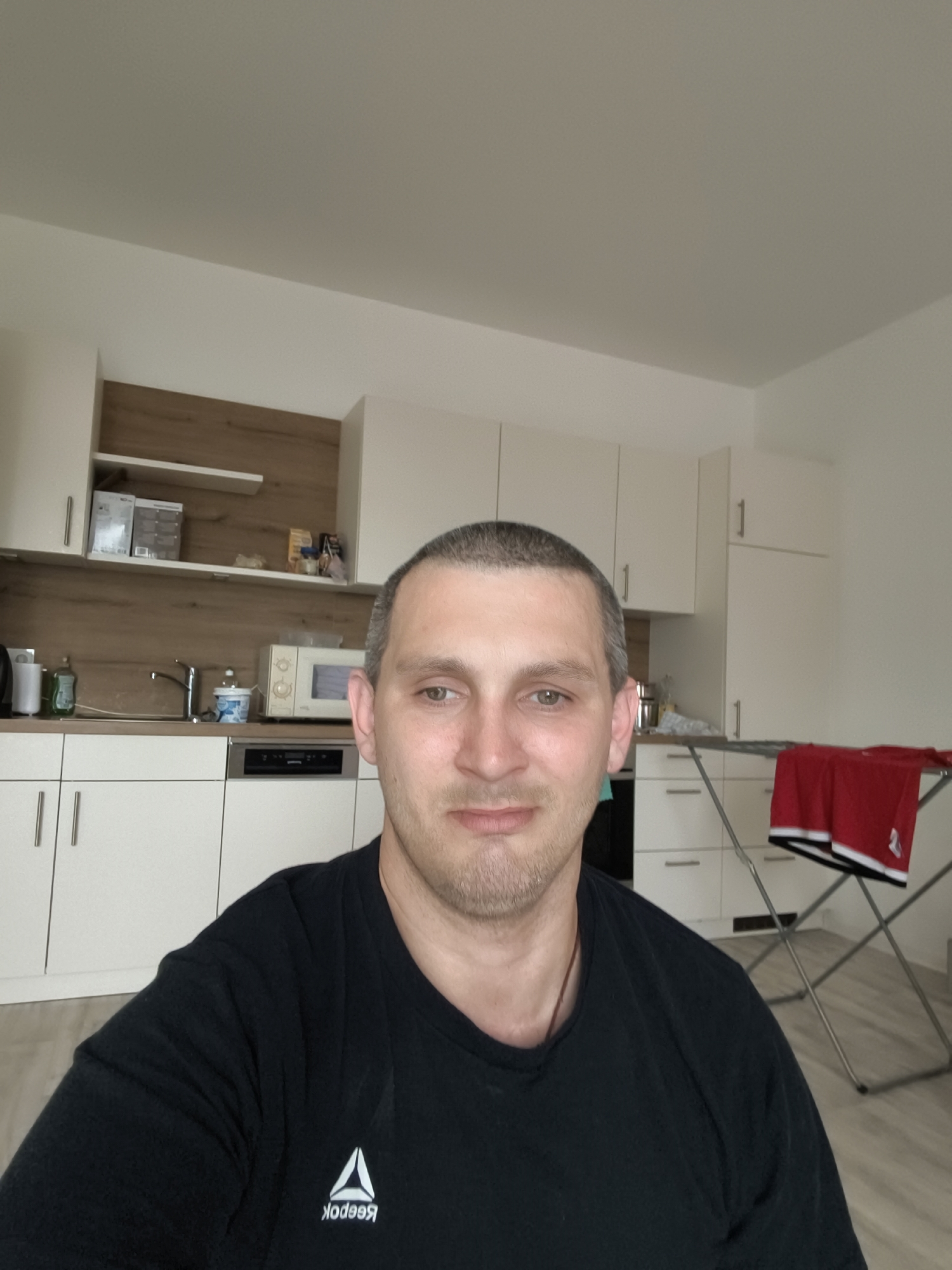 The image is a selfie taken indoors, showing a man with a short haircut and stubble. He is wearing a black t-shirt with a white Reebok logo. Behind him, a kitchen area is visible, featuring white cabinets, a wooden backsplash, a sink with a faucet, a microwave oven, and a clothes drying rack with a red garment. The scene appears to be a residential interior, possibly an apartment, in the city of Wolgast, Germany, given the prompt's context. The lighting is natural, coming from an unseen source, and the overall atmosphere is casual and domestic.