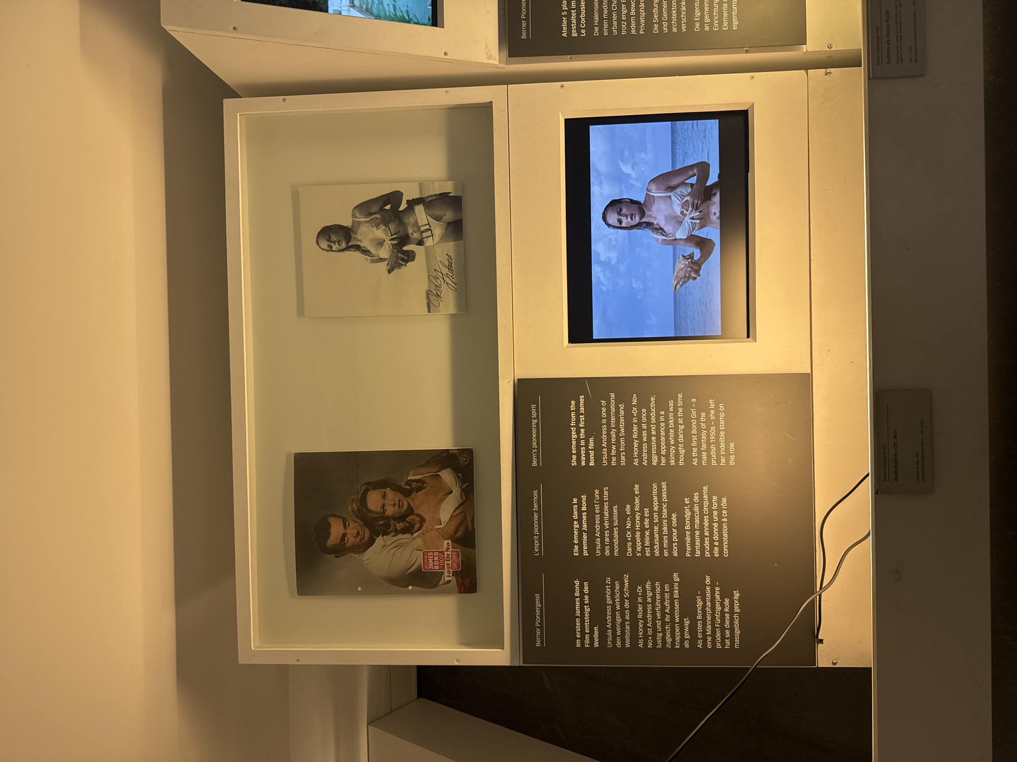 This is a photograph of an exhibit in the Bernisches Historisches Museum in Bern, Switzerland. The exhibit features images and video related to Ursula Andress and her role as Honey Ryder in the James Bond film "Dr. No." In the background, there are two framed images: one is a movie poster featuring Sean Connery and Ursula Andress, and the other is a black and white photograph of Andress. In the foreground, a video is playing on a screen, showing Andress as Honey Ryder emerging from the water. Text panels in German and English provide information about the exhibit.
