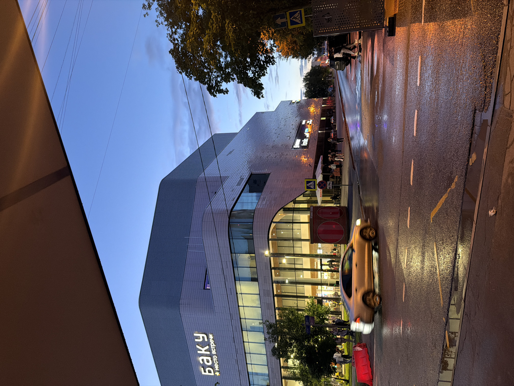 A medium shot captures a street scene in an urban environment at dusk, with a large, modern building dominating the left side of the frame. The building, featuring a gray, corrugated facade and extensive glass windows, has illuminated signage reading "БАКУ" and "* место встречи". A golden sedan is captured in motion, blurring across the wet asphalt road in the foreground. The road surface reflects the ambient light, with visible lane markings and a yellow line. Several people are walking on the sidewalk in front of the building, some under umbrellas or awnings. Trees line the right side of the street, with some illuminated by warm streetlights. The sky is a gradient of deep blue to lighter hues, with some wispy clouds. Traffic signs and a metal fence are also visible on the right. The overall impression is a busy street scene after a rain shower.