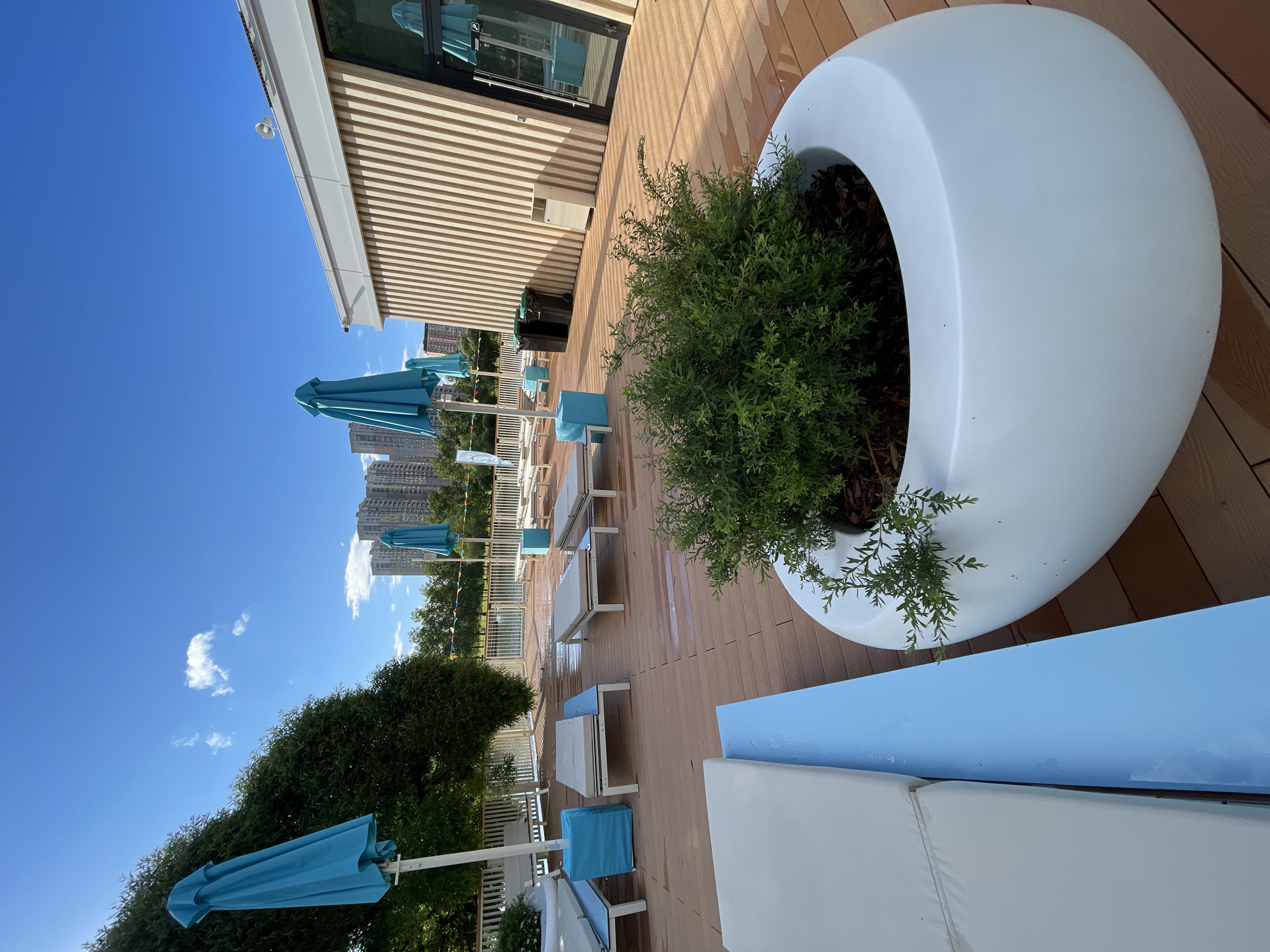This is a ground-level outdoor shot of a modern poolside lounge area in Moscow, Russia. The scene is bathed in bright sunlight under a clear blue sky with a few scattered white clouds.
In the foreground, a large, smooth, white planter pot contains a lush green shrub. To the left, a portion of a light-colored lounge chair is visible, with a vibrant light blue armrest or base. The wooden deck flooring is visible throughout the scene, appearing slightly wet in patches, suggesting recent use or cleaning.
In the mid-ground, several lounge chairs with light-colored cushions are arranged on the deck. Some are empty, while others have blue cubed side tables next to them. Two closed turquoise beach umbrellas stand near some of the loungers. Further back, a modern building with vertical wooden slat siding and large glass windows is visible. Two black trash receptacles and a lighter colored bin are positioned near the building.
In the background, a fence and lush green trees line the area behind the lounge chairs, and a body of water, possibly a pool, is visible. Beyond the trees, several tall, modern apartment buildings characteristic of Moscow's cityscape rise into the sky. The overall impression is one of a well-maintained, contemporary relaxation space. There are no people visible in the frame.