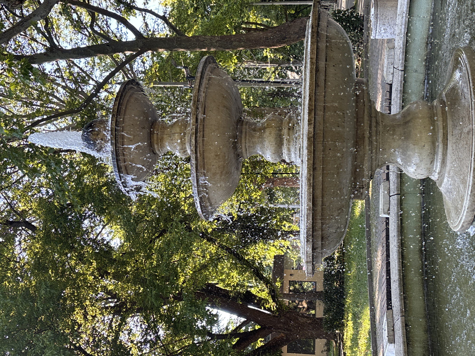 A three-tiered stone fountain is the central focus of the image, positioned in what appears to be a park or public square in Shumen, Bulgaria. Water streams from the top tier, cascading down to the middle and bottom tiers, creating a dynamic spray that is caught mid-air as droplets. The fountain itself is weathered and ornate, with distinct basins and pedestals. Lush green trees with dappled sunlight filtering through their leaves form a verdant backdrop, suggesting a warm, possibly summer day. A paved pathway runs behind the fountain, bordered by grass and some low-lying greenery. In the background, glimpses of buildings and other park elements, such as benches and possibly another smaller stone structure, can be seen. The overall scene conveys a sense of tranquility and nature within an urban setting.