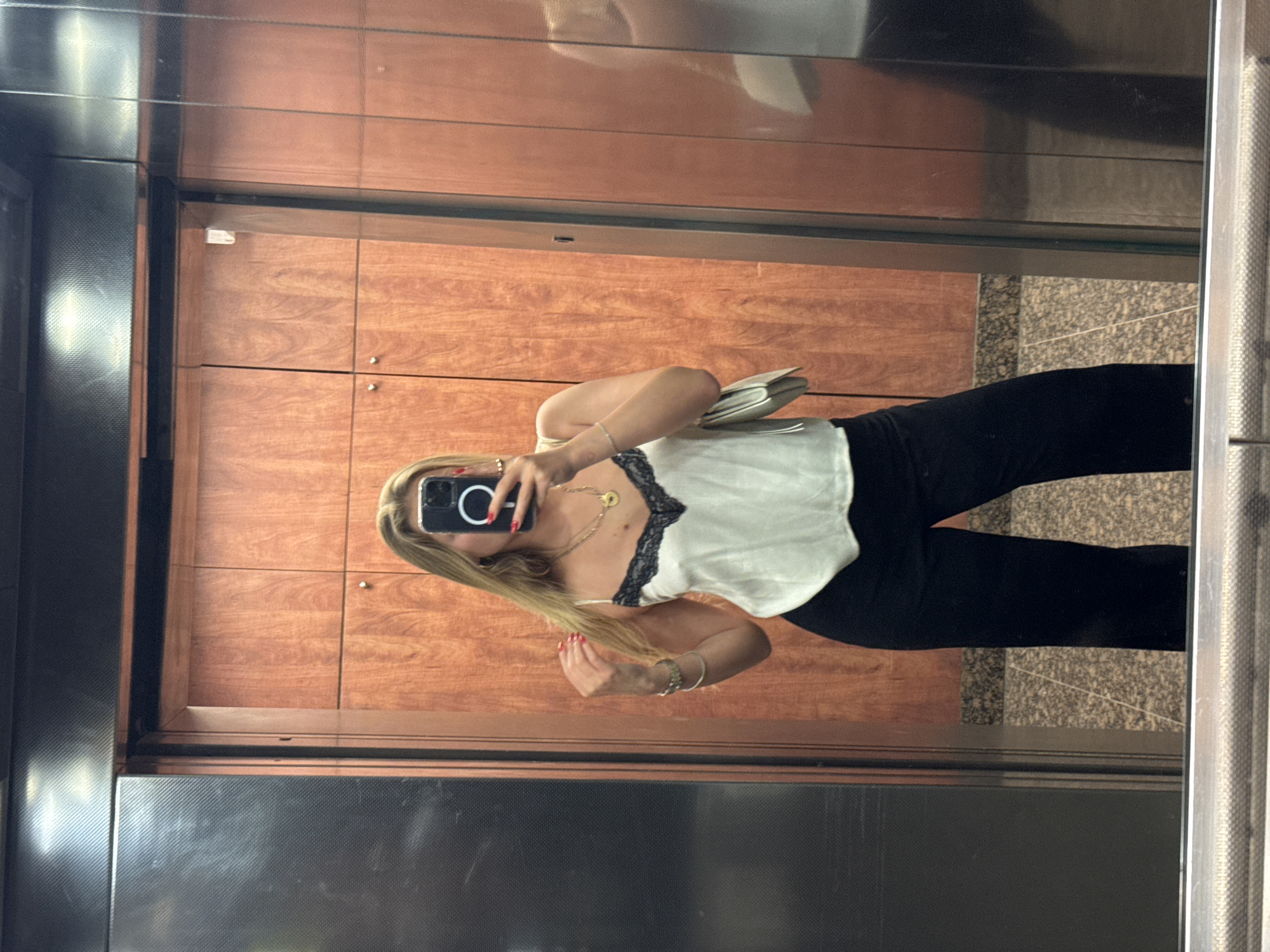 A young woman in black pants and a white top with black lace trim takes a mirror selfie in an elevator. The elevator has wood paneling on one wall and a dark metal finish on another. The floor is tiled. The reflection shows part of what appears to be a hallway or lobby outside the elevator. There is no evidence to suggest the location is specifically Tel-Aviv, Israel.