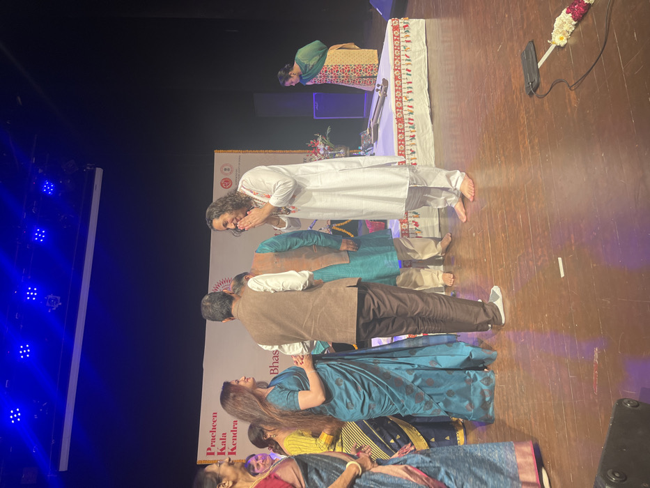 The image shows a group of people on a stage in the Pracheen Kala Kendra in Chandigarh, India (latitude: 30.733, longitude: 76.79). There are five people on the stage: a man in a white shirt with his hands clasped in prayer, a woman in a blue sari, and three men behind them. The man in the white shirt has long hair and is barefoot. The woman is looking at the man in the white shirt. The three men are all wearing casual clothing. The man in the brown jacket is looking down at the woman in the blue sari. The man to his right is looking at the man in the white shirt. There is a man in a teal shirt in the background on the stage. There is a sign on the wall behind the people that reads "Pracheen Kala Kendra" and "Bhas" in Hindi. There is a musical instrument on the stage. There are lights overhead, and the stage is made of wood. There is a woman in a colorful dress in the background who is looking down at the stage. There is a black speaker on the stage and a cord running along the floor to the lower left-hand side of the image. There is a white flower garland on the stage.
