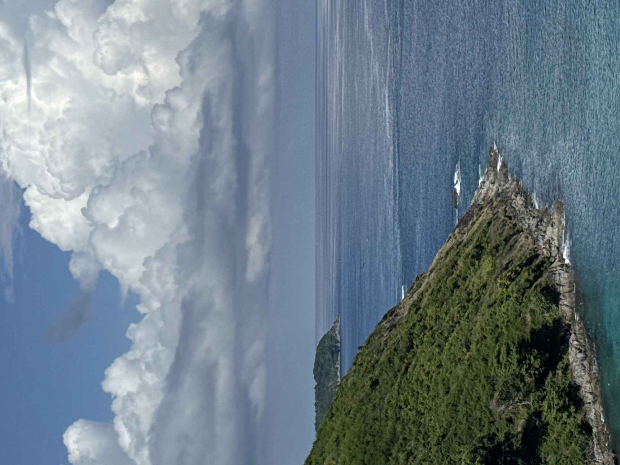 A high-angle, vertical medium shot captures a lush, green island coastline meeting the vast blue ocean under a partly cloudy sky. The foreground is dominated by the steep, verdant slope of an island covered in dense vegetation. Rocky outcrops and the turquoise water are visible along the base of the island. The sea stretches out to the horizon, where it meets a hazy blue sky. Puffy, white cumulus clouds drift across the sky, interspersed with darker, more scattered clouds. A distant, smaller island with green foliage is visible on the left side of the frame, further out in the ocean. The scene depicts a natural, tropical landscape with no people or man-made structures clearly visible.