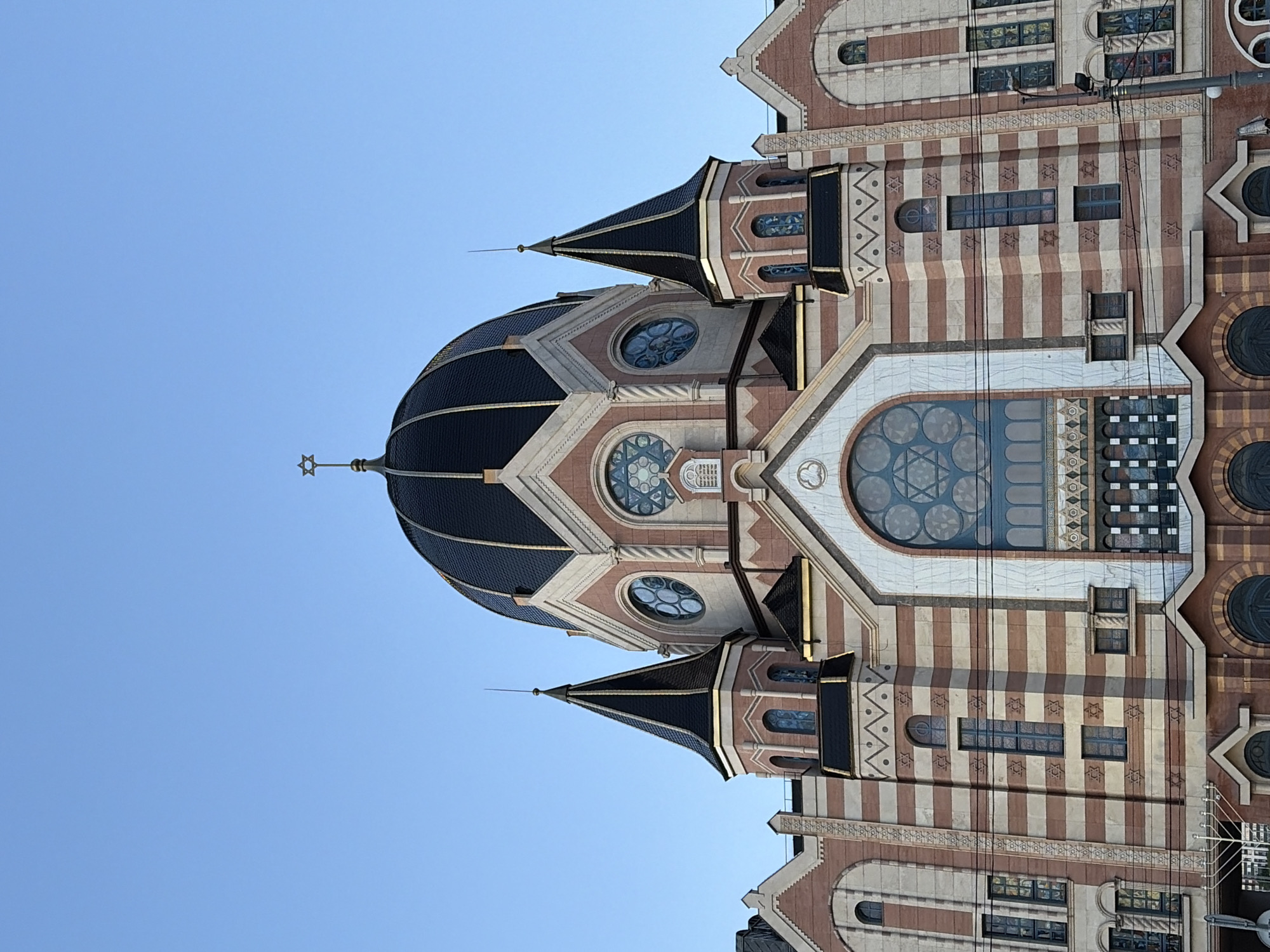 The image is an upward-facing shot of a large, ornate building, likely a synagogue, against a clear blue sky. The building features a prominent dark dome topped with a Star of David, and two smaller, dark, pointed turrets on either side. The facade is constructed with alternating bands of light and dark brick, accented with light-colored stone or stucco. Several windows are visible, including large arched windows with intricate stained glass designs, and smaller circular windows. The building's architecture appears to be a blend of styles, possibly Moorish Revival or Neo-Byzantine, given the dome and decorative elements. There are no people or vehicles visible in the image, and the focus is solely on the exterior of the building. The environment is clear and bright, suggesting it is daytime with good weather. The building is situated in an urban setting, as indicated by the presence of utility poles and wires in the foreground.