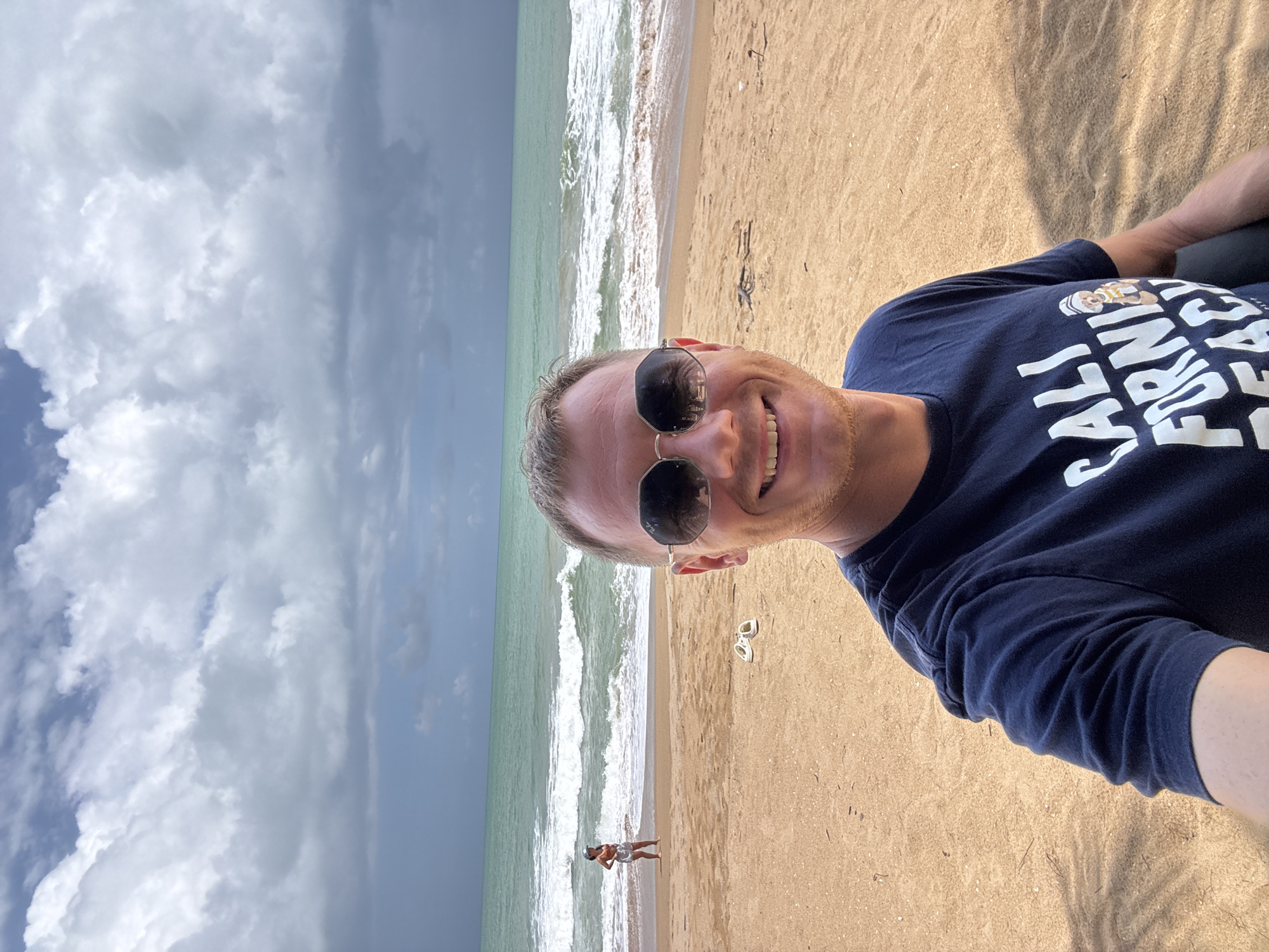 A selfie taken on a beach shows a smiling man with short brown hair wearing sunglasses and a navy blue t-shirt with white lettering that reads "CALI FORNI BEACH" and a small cartoon bear. He is positioned in the foreground. Behind him, the beach is a light tan color. Further back, a woman in an orange top and white shorts stands near the water's edge, looking out at the sea. White waves are breaking on the shore. The ocean is a greenish-blue. The sky is filled with dramatic, dark gray and white clouds. In the middle ground on the sand, a pair of white flip-flops are visible. The scene is likely in the city of Ban Khuk Khak, Thailand, given the tropical beach environment.