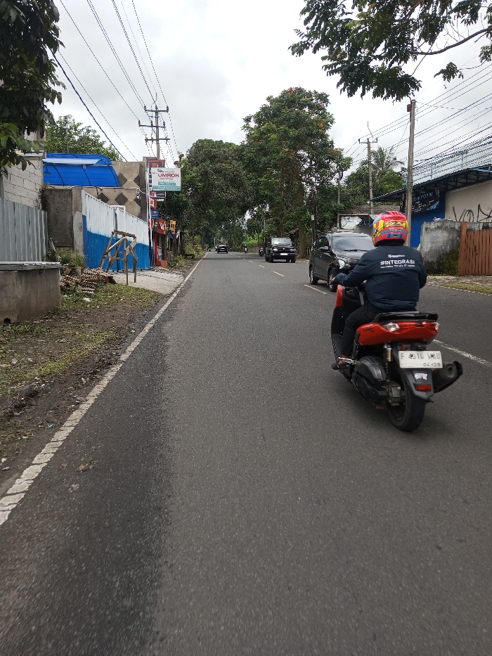 An individual on a red and black scooter drives away from the viewer on an asphalt road in Serpang Shalli, Bhutan, under an overcast sky. The rider wears a red helmet with yellow and black markings and a dark blue jacket with "INTEGRASI" in white text on the back. The scooter's rear license plate displays "F 4316 HAU" with "04.28" below. To the left, a dirt shoulder borders a building featuring a blue corrugated roof extension, white and blue panels, and a commercial sign for "UMROH". Utility poles with overhead power lines are present. In the opposing lane, a black car and other distant vehicles are visible. The environment features trees with green foliage lining both sides of the road, and a concrete wall with graffiti alongside a brown wooden gate is observed on the right.