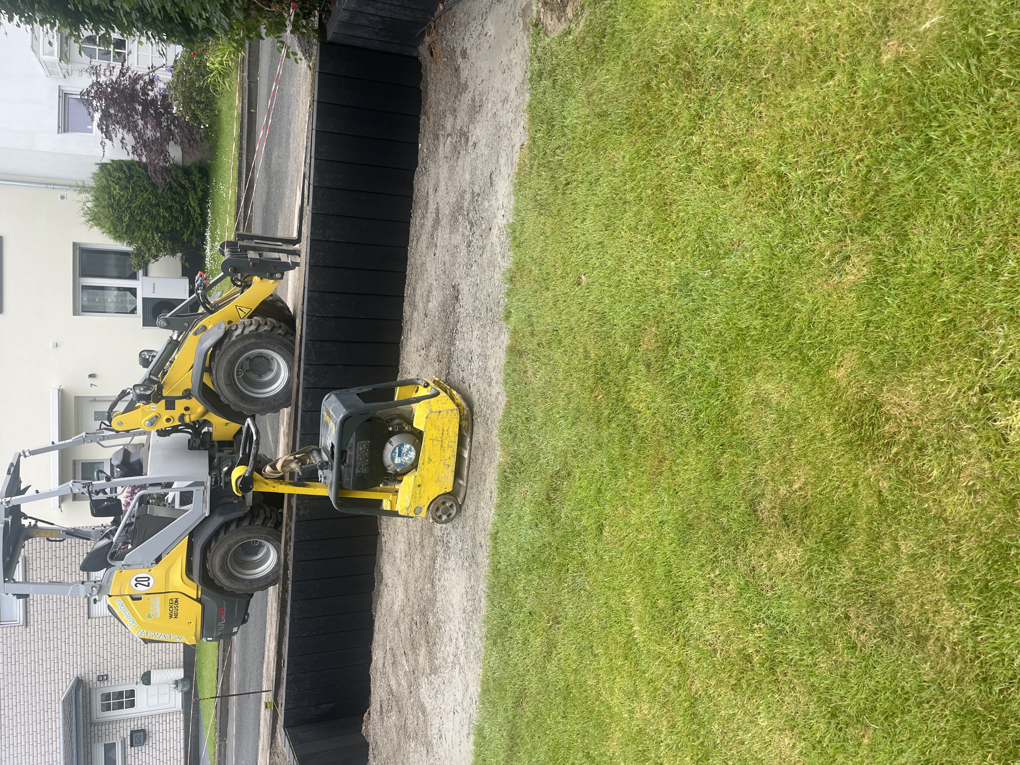 The image depicts an outdoor scene showcasing a landscaping or construction project in a residential area, under bright daylight. The central focus is a prepared strip of bare earth, bordered by a dark retaining wall and a lush green lawn.
On the left side of the frame, a yellow compact wheel loader, identified by the visible text "KASIRATI Gartenbau" and "WACKER NEUSON VL 20" along with the number "20", is positioned on a paved surface, likely a driveway or street. Behind this vehicle, residential buildings are visible, including a white house with various windows and a distinct white entrance, and another brick-faced house further back. Green foliage, comprising trees and a dark red-leaved bush, adds natural elements to the background. A red and white striped barrier tape is stretched across the upper left, demarcating a work area.
The middle section of the image is dominated by a dark black retaining wall, constructed from horizontal planks. Adjacent to this wall is a long, light brown strip of bare earth or sand, which appears to have been leveled and possibly compacted. A yellow vibratory plate compactor, with the brand "WACKER NEUSON" partially visible, rests on this strip, indicating active ground preparation work.
To the right, a wide expanse of vibrant green lawn stretches across the rest of the image, showcasing healthy, cut grass. The distinct boundary between the prepared earth and the existing lawn suggests an ongoing transformation or development of the outdoor space. The scene captures a moment of construction activity in a suburban setting.