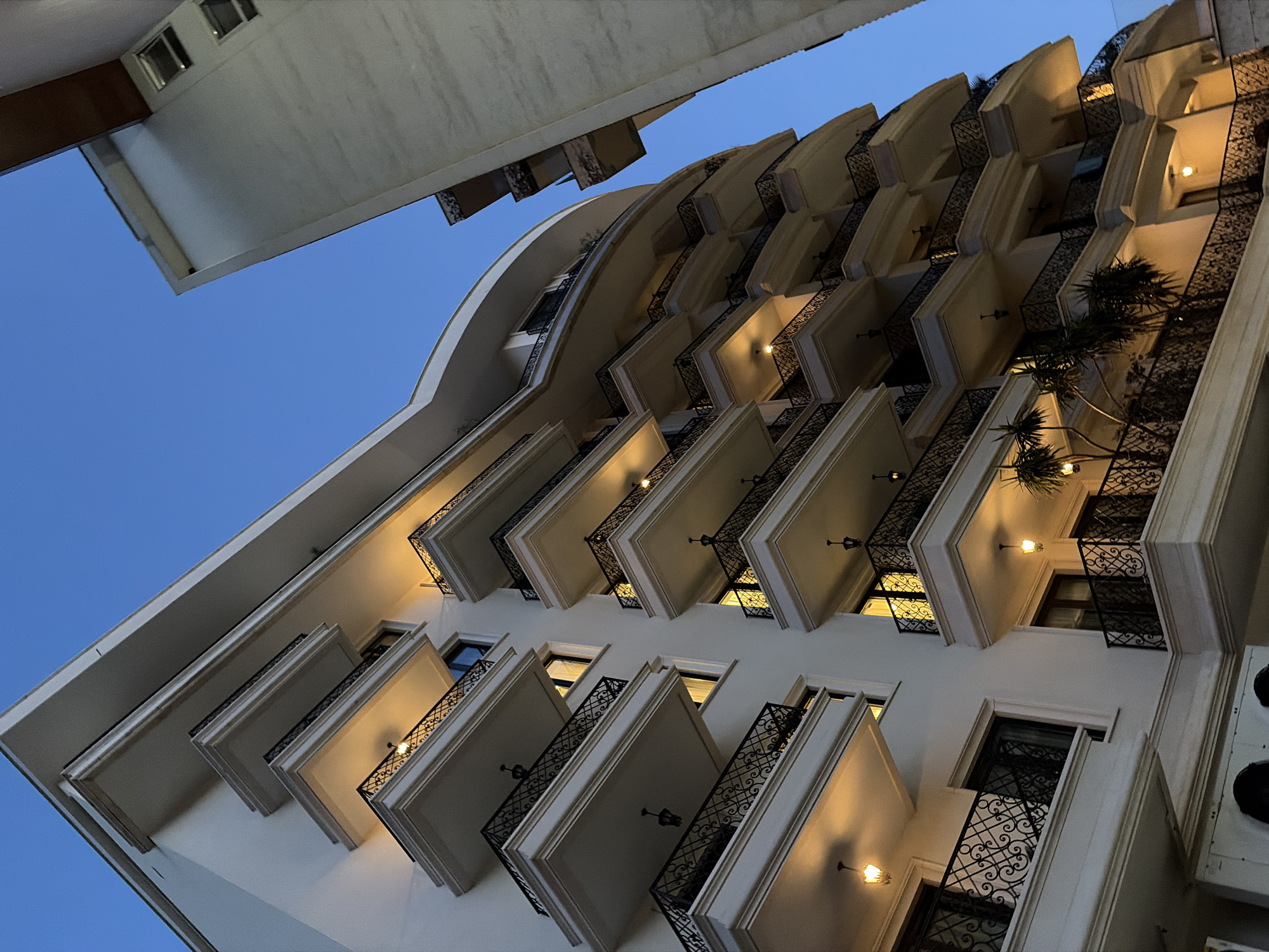 The image shows a modern, multi-story apartment building with numerous balconies, set against a clear, twilight sky. The building has a curved facade on the left and a more angular structure on the right, with the sky filling the upper portion of the frame. Each balcony is adorned with ornate black railings and illuminated by warm, yellow lights. Some balconies feature small trees or potted plants. The lower section of the building reveals white air conditioning units. Another building, partially visible on the right, appears to be under construction or renovation, with a lighter colored facade and some visible windows. The scene is likely in Budva, Montenegro, given the architectural style and the prevalence of such buildings in coastal Montenegrin cities. There are no people visible in the image.