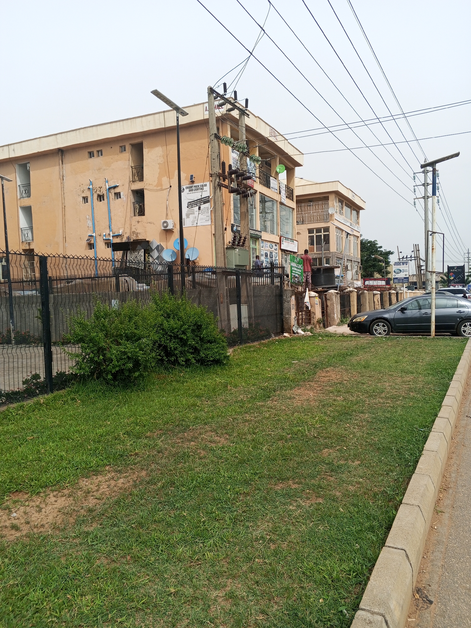 The image depicts a street scene in Abuja, Nigeria. In the foreground is a grassy area separated from the street by a concrete curb. Behind the grass is a black metal fence, and behind the fence is a two-story building with a peach-colored exterior. The building appears to house several businesses, indicated by signs on the exterior. A utility pole with electrical equipment stands adjacent to the building. A dark-colored car is parked along the curb. A man in a red shirt is standing on a structure adjacent to the fence, seemingly working on something. Two other people are visible near the fence. Overhead are numerous electrical wires. The sky is overcast.