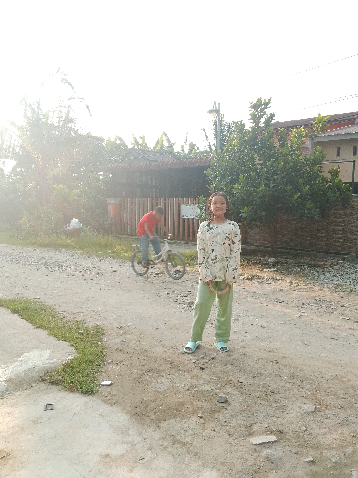 In Hamparan Perak, Indonesia, a young girl stands on a dirt road, smiling at the camera. She is wearing light green pajama pants and a long-sleeved white shirt with a playful pattern of cartoon animals. On her feet are light blue sandals. She is holding a round, beige object with her right hand. Behind her, a young boy in a red shirt and blue jeans rides a bicycle along the same road, his motion captured in a blur. The background features a fence, trees, and buildings, suggesting a residential area. The sun is bright, casting a warm glow over the scene. The road is unpaved, with scattered stones and patches of grass growing along the edges.