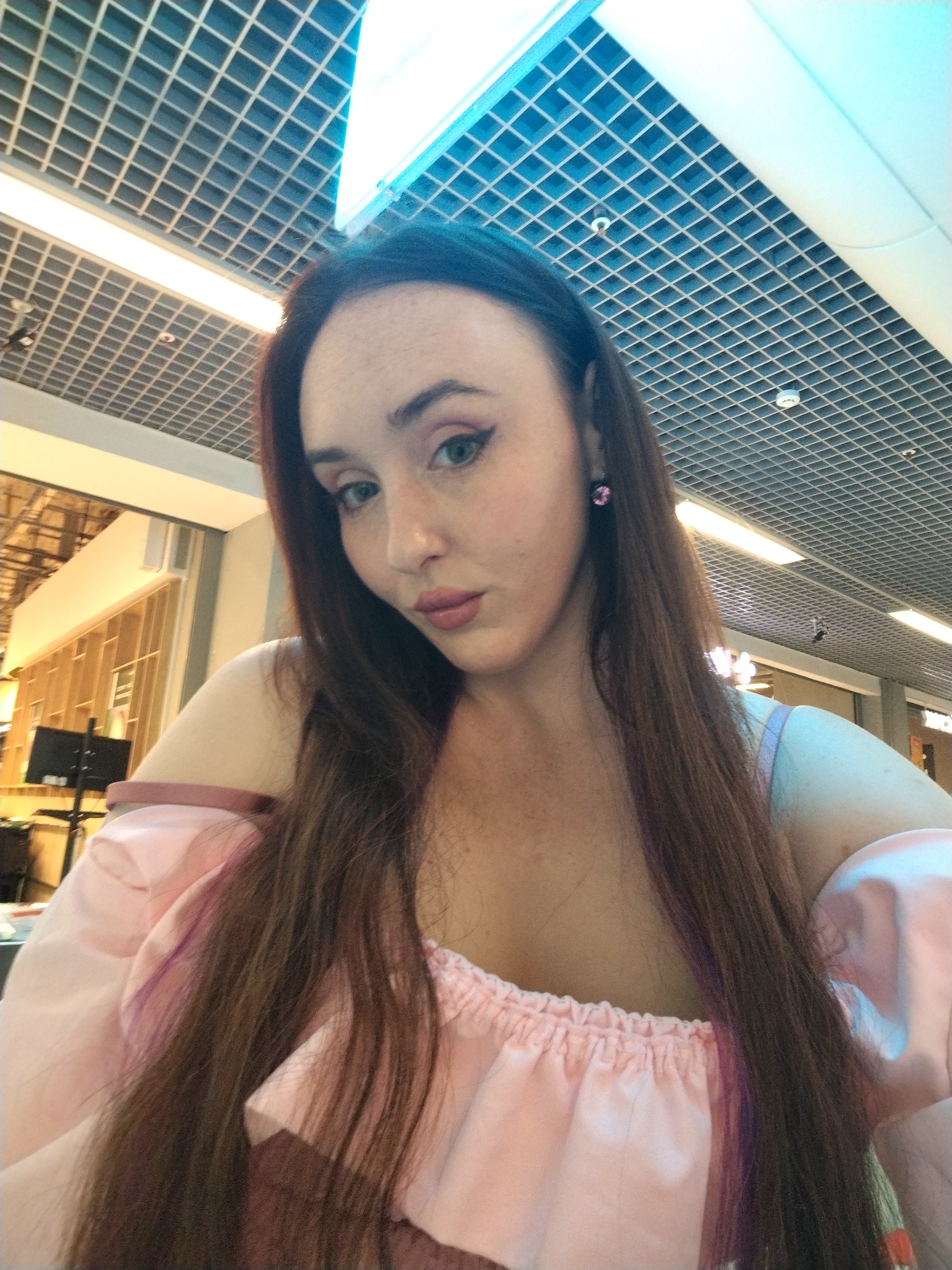 A young woman with long, dark brown hair and fair skin with freckles is captured in a close-up, angled selfie. She has green eyes, a pink-toned lipstick, and is wearing a pink off-the-shoulder top with ruffled detailing. A pink stud earring is visible in her left ear. The background appears to be an indoor public space with a grid-patterned ceiling and bright overhead lighting. The overall lighting suggests a well-lit interior environment. There is no indication of this specific location being within the city of Izhevsk, Russia.