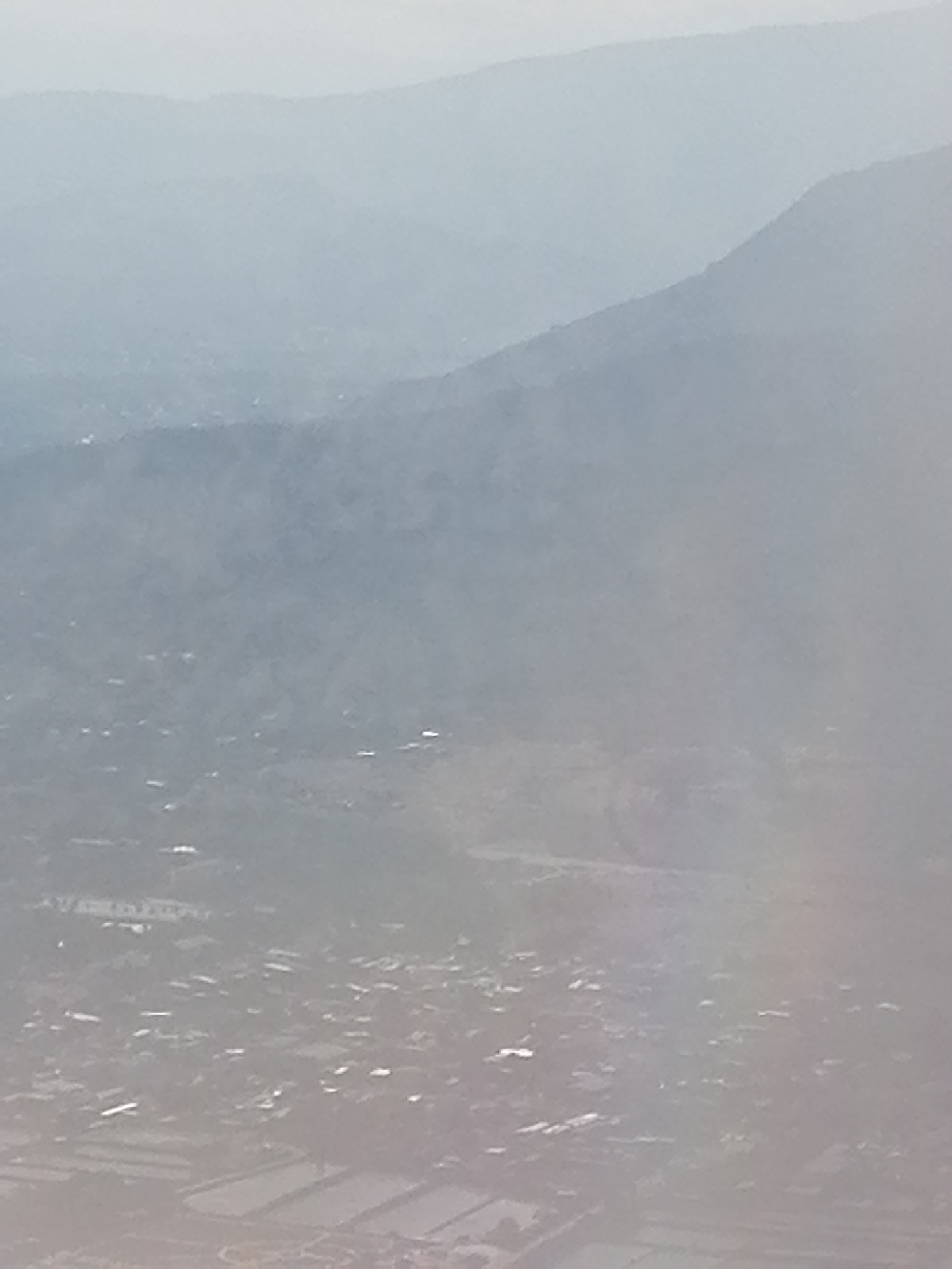 The image depicts a hazy, aerial view of a landscape featuring mountains in the background and a settlement in the foreground. The mountains exhibit a gradient of blue-gray hues, indicating varying distances and atmospheric perspective. The settlement is comprised of numerous small structures and fields, discernible but blurred due to the atmospheric conditions. The general environment is overcast and indistinct.