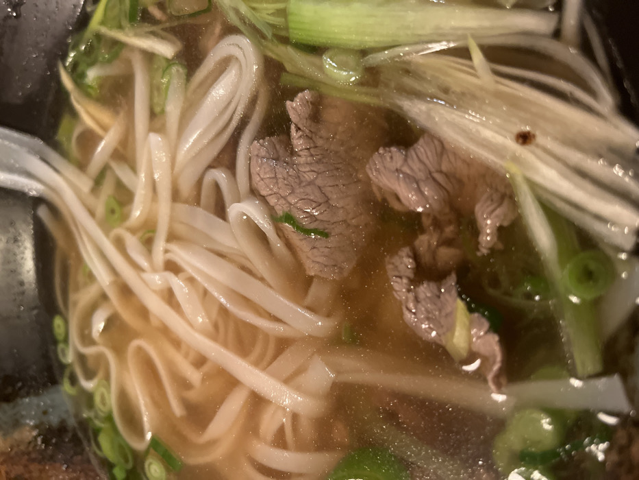 A close-up shot captures a bowl of Vietnamese Pho. Flat rice noodles intertwine with thin slices of beef in a clear, light broth. Chopped green onions are scattered throughout, adding a fresh element. The image focuses on the textures and colors within the bowl, highlighting the noodles' creamy white hues against the broth's golden transparency. The beef appears partially cooked, revealing its grain.