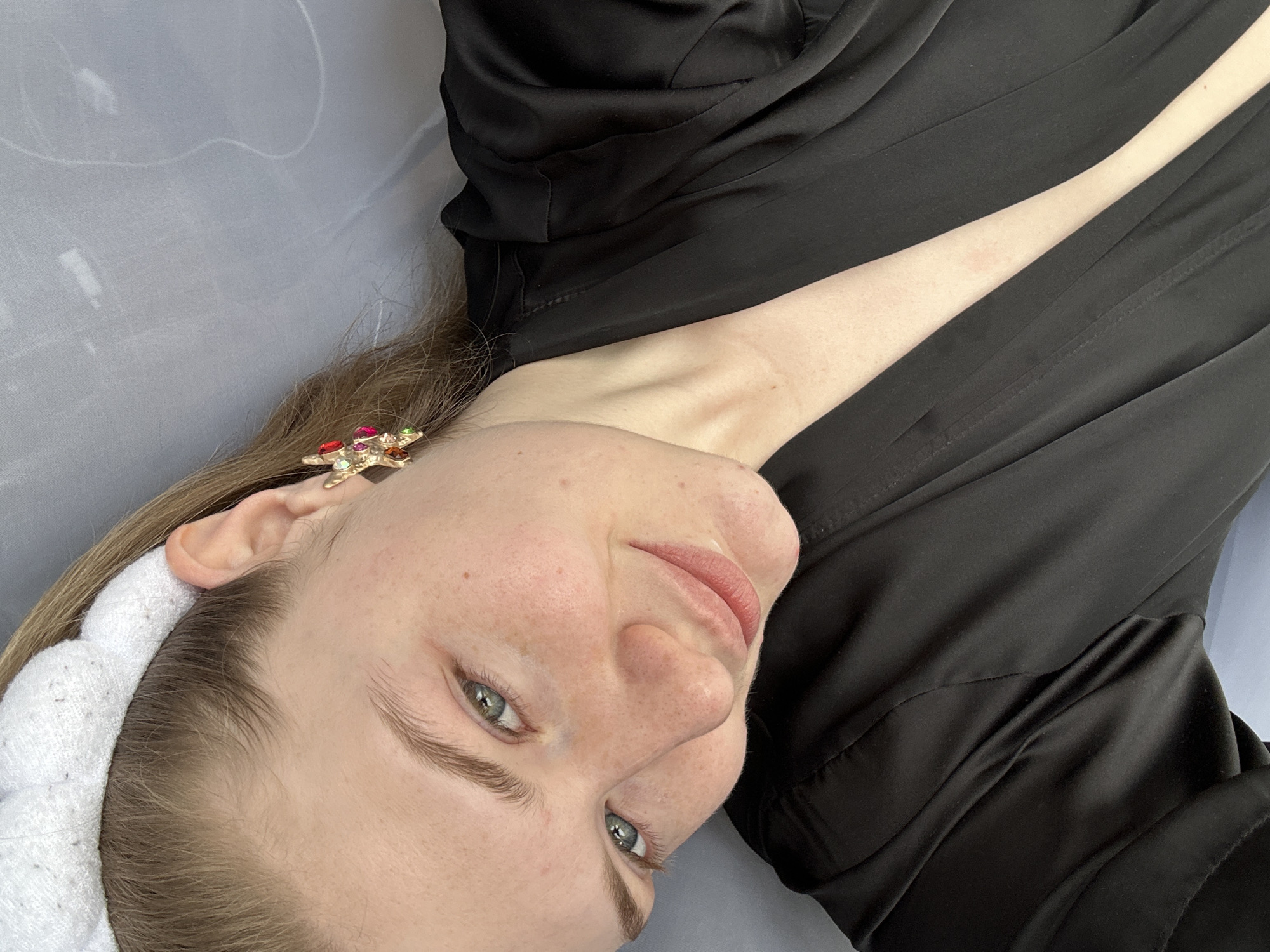 A woman lies on her side on a gray surface. She is wearing a black garment and a white headband. A colorful star-shaped earring adorns her ear. Her eyes are open, and she is looking at the camera. The location is not specified.