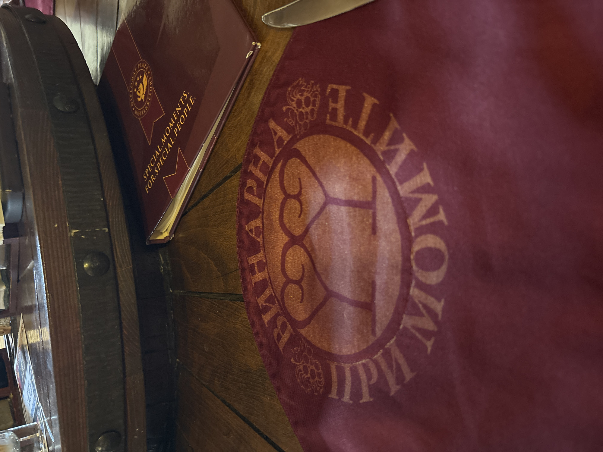 The image shows a close-up of a table setting at a restaurant in Lukovit, Bulgaria. A dark wooden table with a riveted edge is in the background. On the table, there is a maroon menu with gold lettering that reads "SPECIAL MOMENTS. FOR SPECIAL PEOPLE." A circular logo with stars and the word "DIPLOMAT" is visible on the menu. In the foreground, a maroon cloth placemat with a gold circular emblem is laid out. The emblem features a stylized representation of two wine glasses and what appears to be grapes or a vine. The text within the circle on the placemat is in Cyrillic script, possibly reading "ВИHАРНА" and "ПРM MOMHTE". A silver knife is partially visible on the right side of the frame. The environment suggests an indoor dining establishment.