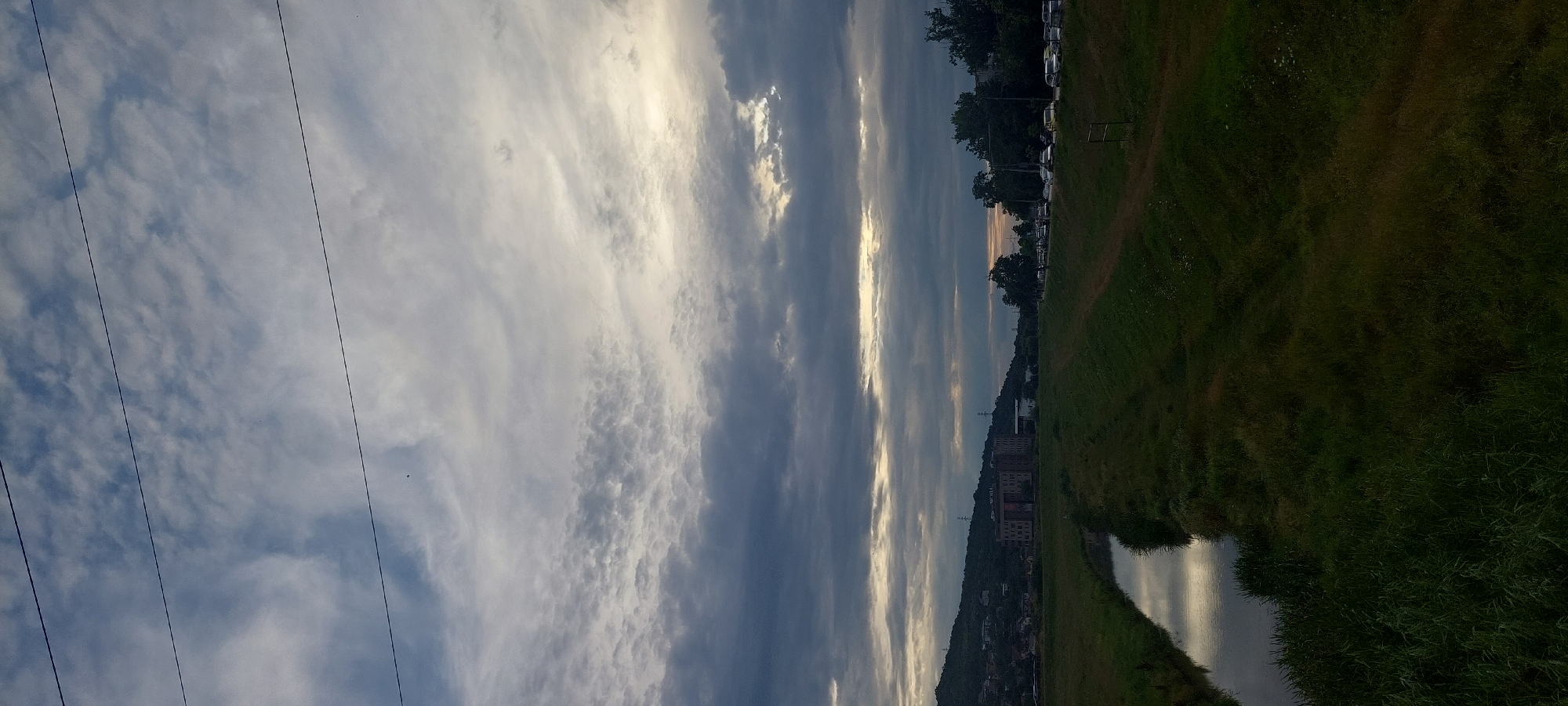 The image depicts a somewhat overcast sky with layered clouds, ranging from dark grey to lighter white, with a sliver of bright light breaking through towards the horizon on the right side. Three thin, dark power lines are stretched horizontally across the upper portion of the frame. In the middle ground, a small body of water, likely a canal or a narrow river, reflects the sky. The banks of this waterway are covered in green grass, with a darker, rougher embankment on the right.
Beyond the water and grassy slopes, a town or city landscape is visible on the left, nestled against a hill. Several buildings, some appearing to be apartment blocks or industrial structures, are situated on the hillside, with houses scattered amongst them. A larger, darker building with multiple floors and visible windows stands prominently. Further to the right, a line of trees borders a road where several cars are parked, suggesting an urban or suburban setting. The overall scene appears to be captured during the daytime, possibly late afternoon or early evening, given the lighting conditions.
Based on the visual cues of the architecture and the landscape, this scene is located in the city of Iași, Romania. The characteristic hills, building styles, and the presence of the canal strongly suggest this location, likely in an area adjacent to the Copou hill or along the Bahlui river.