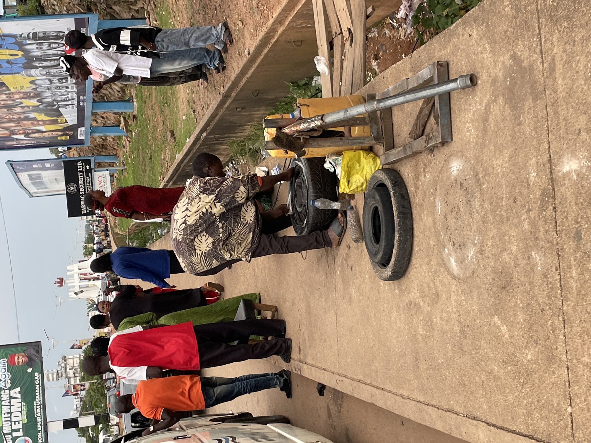 The image depicts a street scene in Jos, Nigeria, possibly near a major road intersection. In the foreground, a man in a patterned shirt and dark pants is working on a tire using a metal tool. Several tires are visible on the ground, along with a yellow jerrycan and a yellow plastic bag. A white van is parked on the left side of the frame.
In the background, several people are standing on the sidewalk. One man wears a red shirt and black pants, another is in an orange shirt, and a woman in a black dress and headscarf is also present. Further in the background, there are billboards, traffic, and a pedestrian bridge with a distinctive crown-like structure. The environment appears to be urban, with a mix of commercial signage and street infrastructure. The sky is overcast.