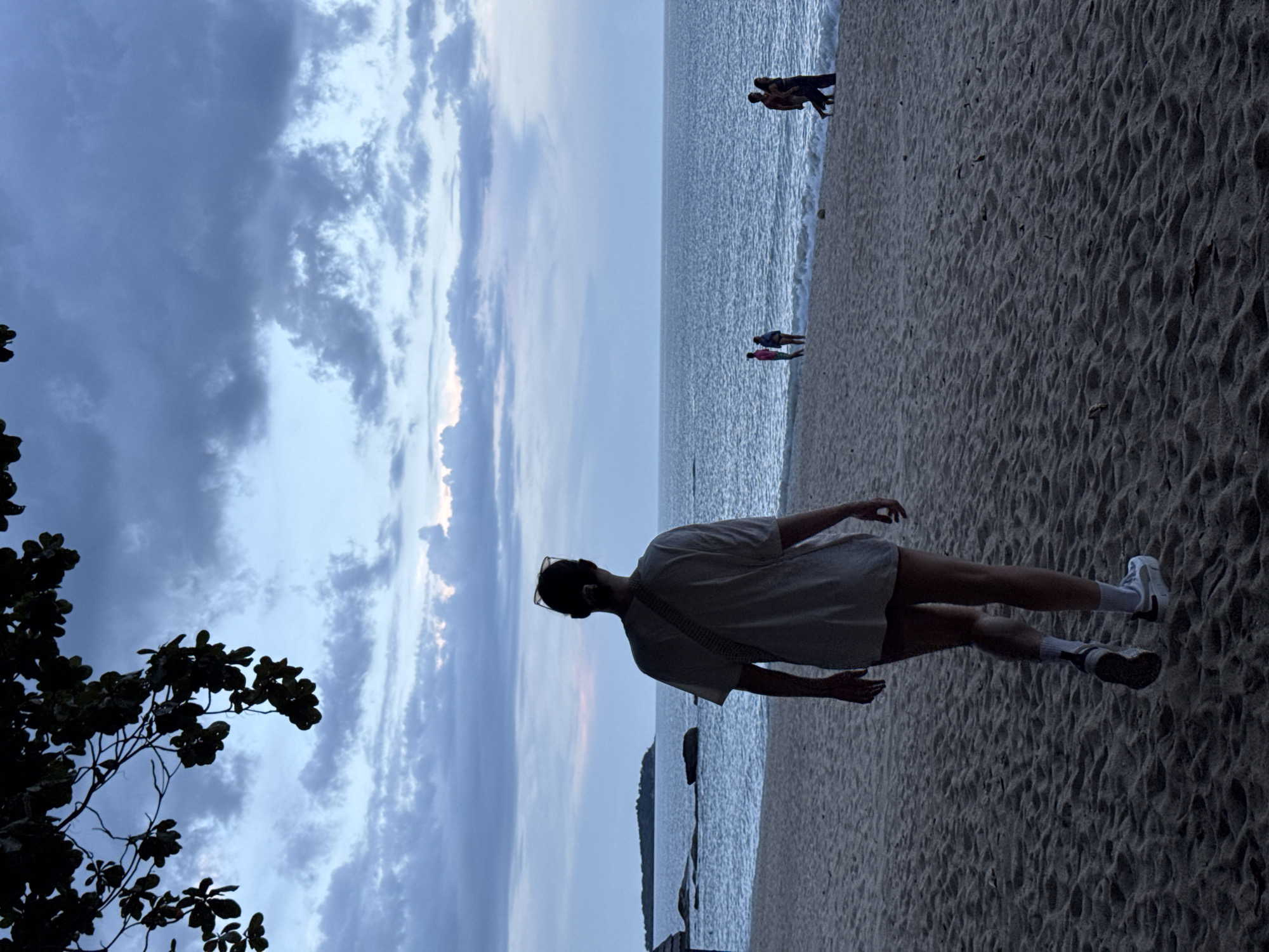 This media file captures a person standing on a sandy beach in Baan Chaweng Noi, Thailand. The image is oriented vertically, with the person positioned in the foreground, facing away from the camera and towards the ocean. They are wearing a light-colored t-shirt, shorts, white socks, and sneakers. A bag is slung across their shoulder. Sunglasses are perched on their head.
The scene includes the ocean in the background, with gentle waves lapping at the shore. The water appears textured and reflects the overcast sky. Several other individuals are visible in the distance, some near the water's edge and others further up the beach. A branch with dark leaves from a tree is silhouetted in the lower left corner of the frame, adding a natural element to the composition.
The sky is filled with dramatic clouds, suggesting either early morning or late evening. The light is muted, indicating a time of day with diffused sunlight, possibly before sunrise or after sunset, or a cloudy day. The overall mood appears serene and contemplative, with the vastness of the ocean and sky creating a sense of scale. The sandy texture of the beach is prominent, showing the intricate patterns left by the receding waves. No text is visible in the image.