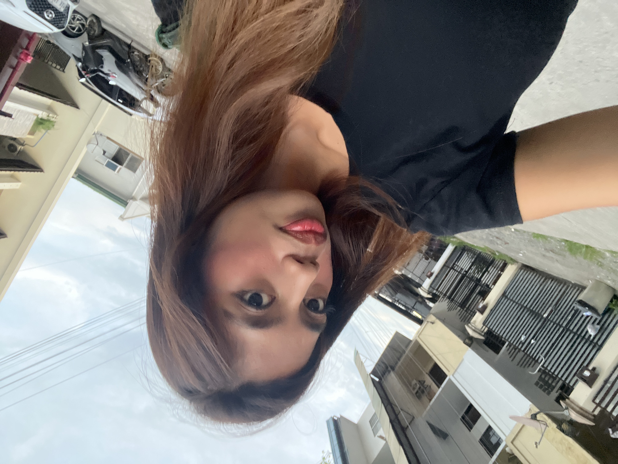 A selfie-style image features a woman in an outdoor urban setting. The image is originally rotated 90 degrees counter-clockwise, showing the woman's head tilted.
**Subjects:**
The primary subject is a young adult woman with long, wavy brown hair, parted on her left side, which cascades over her shoulders and frames her face. She is wearing a simple black t-shirt or top. Her face is prominently featured, with noticeable makeup including red-pink lipstick, blush on her cheeks, and dark eyeliner/mascara. She is looking directly into the camera with a neutral expression, her lips slightly parted.
**Scene and Setting:**
The background reveals a street or residential area, consistent with an urban or suburban environment. On the left side of the frame, behind the woman's head, an overcast sky is visible with thin electrical wires stretching across. Below the wires, parts of light-colored buildings with windows and an air conditioning unit are seen. On the right side, there are more buildings, some with black metal fences or gates, suggesting residential or commercial properties. A white SUV is parked on what appears to be a driveway or street, with a partial license plate displaying "XFV 717." Another darker vehicle is partially visible behind it. Some green foliage is also present on the ground.
**Activity or Event:**
The woman appears to be taking a self-portrait or being photographed, given her direct gaze at the camera.
**Notable Details:**
The scene is set during the daytime, under an overcast sky. The overall environment, including the architecture and vehicles, is typical of an urban area in Southeast Asia, aligning with the provided location context of Davao City, Philippines. The woman's expression is neutral, perhaps slightly pouty.
**Visible Text:**
The partial license plate on the white SUV clearly shows the alphanumeric sequence "XFV 717".