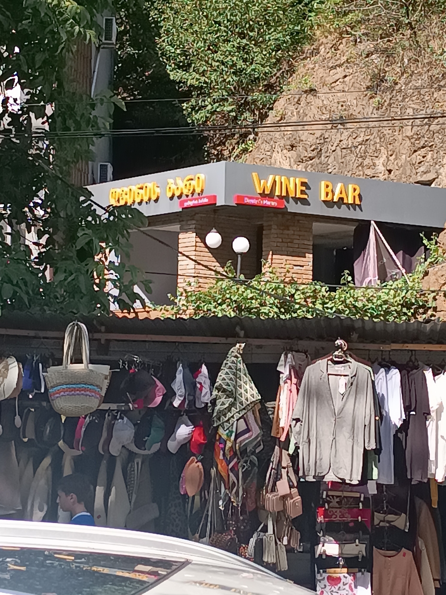 The image depicts a street scene in Borjomi, Georgia. A prominent sign in yellow lettering with a Georgian inscription above it reads "WINE BAR". Below the "WINE BAR" text, in a red rectangular box, is smaller text that reads "Dimitri's Marani". The establishment appears to be built into a rocky hillside, with dense green foliage surrounding the upper portions of the building. In front of the wine bar, a stall or market displays a variety of clothing items and accessories, including jackets, shirts, bags, and hats, hanging on racks. A young boy, wearing a blue and white shirt, is visible looking to the left behind the display of clothing. The foreground shows the roof of a vehicle. The lighting suggests it is daytime, with shadows indicating sunlight.