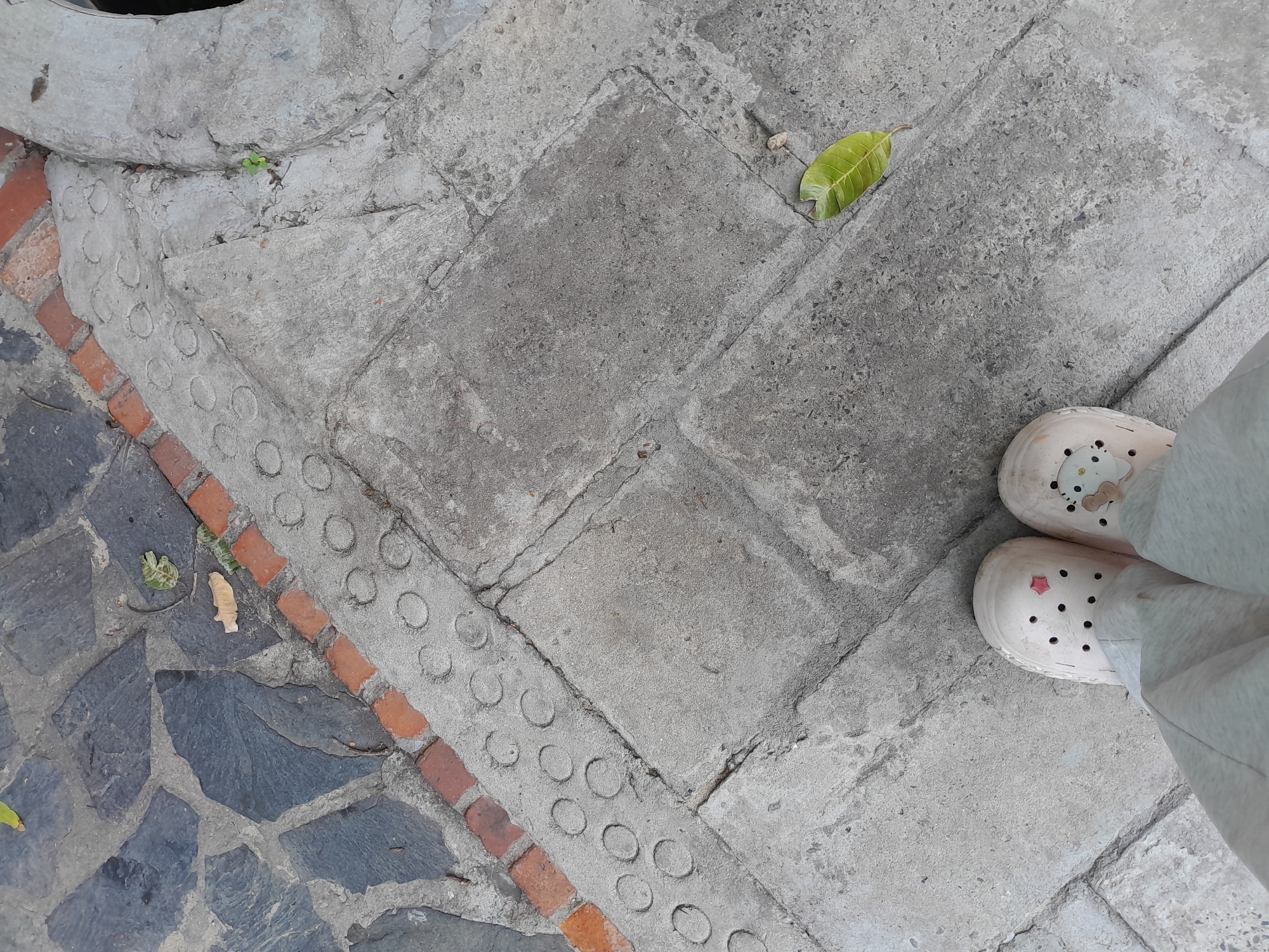 The image shows a ground-level view of a pedestrian walkway in Phan Thiet Ward, Vietnam. The walkway is constructed with a variety of materials including large, irregular grey stone slabs, smaller red brick edging, and concrete sections with raised circular patterns, likely tactile paving for the visually impaired. A light-colored shoe with polka dots and a small pink star charm is visible at the bottom of the frame, along with light grey pants. There are fallen leaves on the ground, one prominently green and veined, and other organic debris. The overall impression is of an outdoor urban environment with worn surfaces.