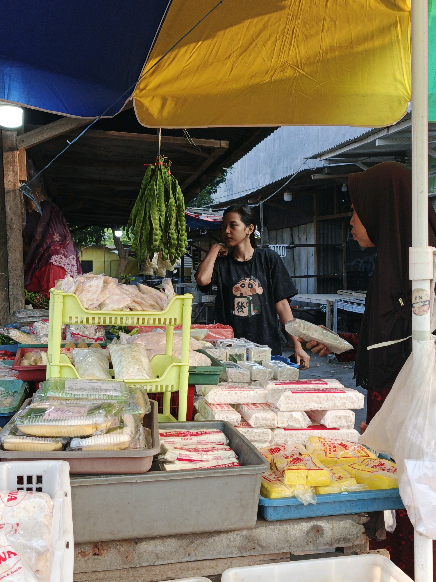 This image depicts an outdoor market stall, likely in Kalisampurno, Indonesia, with a focus on packaged food items. Two women are present. One, wearing a black t-shirt with a cartoon character, stands behind a display of green, pod-like vegetables hanging from the stall's structure. The other woman, wearing a dark hijab and a black outer garment, is shown from the side, holding a package of food. The stall is laden with various food products, including what appears to be tempeh, tofu, and other packaged goods, predominantly white and yellow in color, arranged on shelves and in trays. The market environment is suggested by the rustic wooden structures and roofing, along with a blue and yellow tarp overhead. The overall scene is one of commerce, with the women engaging in the transaction or preparation of goods for sale.