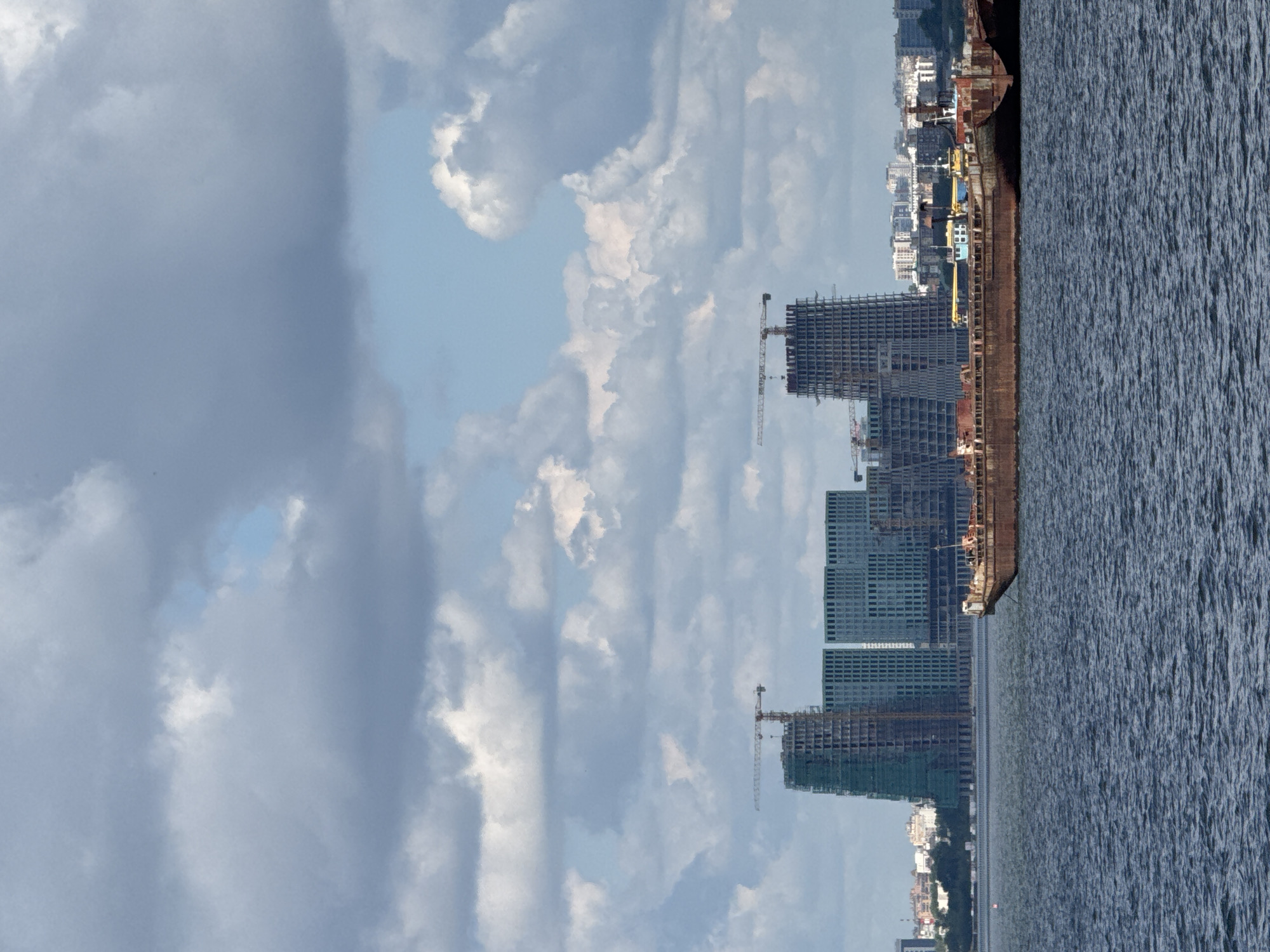 The image shows a body of water with several large, rusty barges in the foreground. In the background, a city skyline is visible, dominated by several tall buildings under construction, some with cranes. The sky is filled with dramatic, heavy clouds. This scene is likely the Volga River in Kazan, Russia, near the city's growing business district, which features many modern high-rise developments. There are no people visible in the image, and the barges appear to be stationary.
