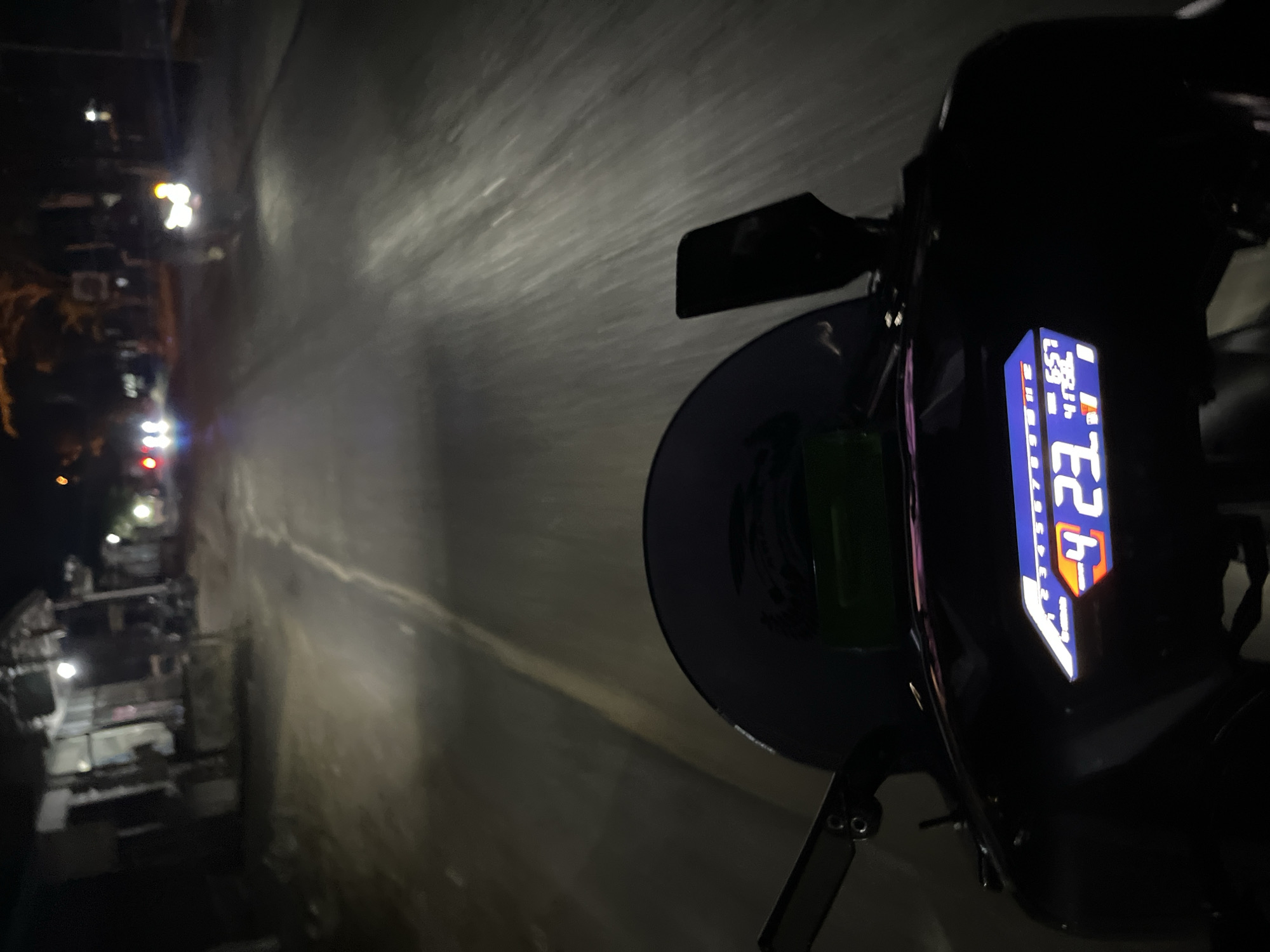 The image shows a first-person perspective of someone riding a motorcycle at night on a road in Pecangaan, Indonesia. The road appears to be paved and has a center line. There are streetlights visible in the distance, as well as headlights from oncoming vehicles. On the left side of the road, there are some buildings, possibly shops or houses, with lights on inside. The motorcycle's digital dashboard is visible in the foreground, displaying speed, gear, and possibly other information. The dashboard shows the numbers "4", "2", "3", and "657". The environment is dark, with the only significant light sources being artificial lights from the road and buildings.